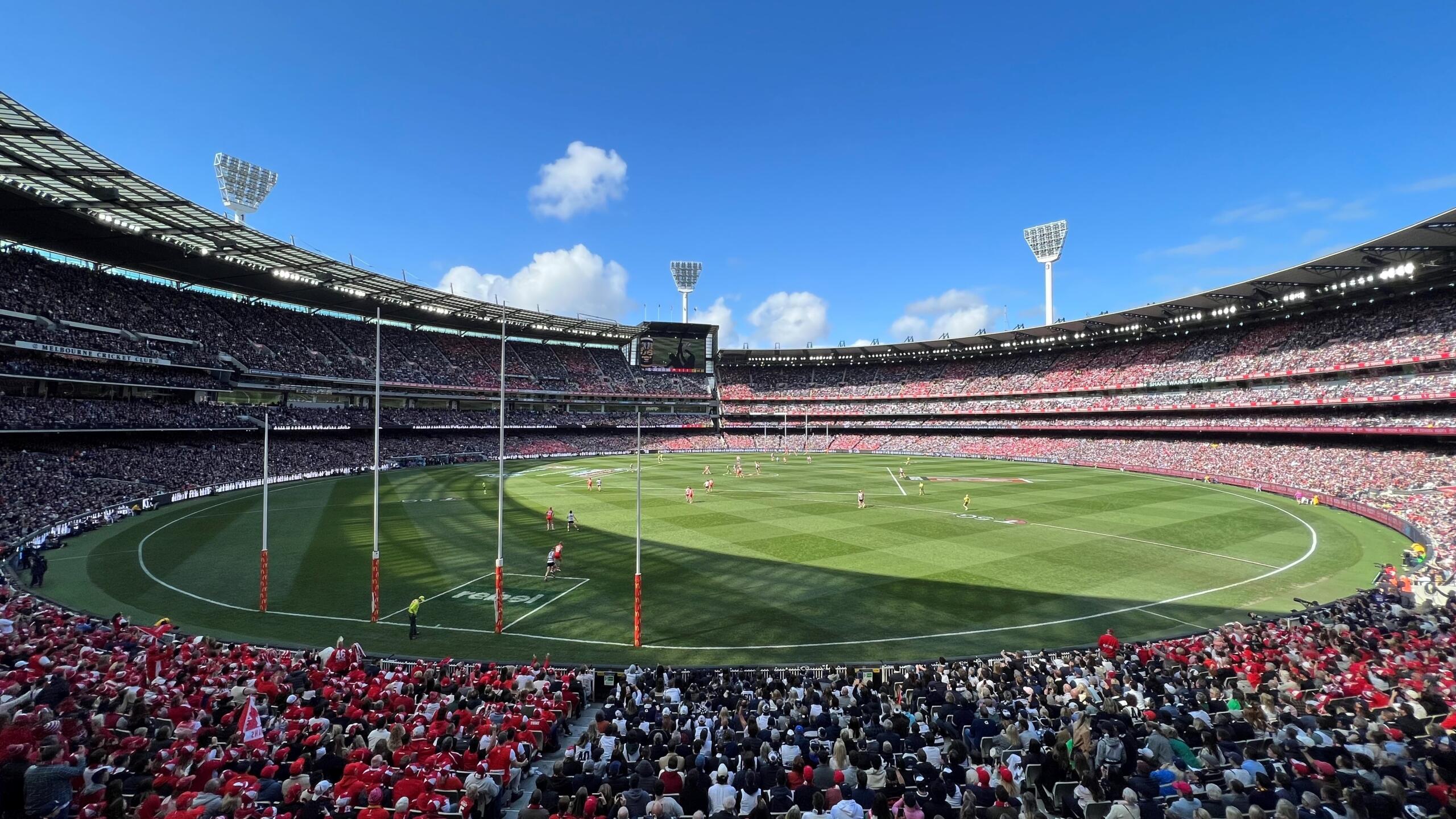 A panoramic view of a packed stadium hosting an AFL final match, with players on the field and fans in red and black attire.