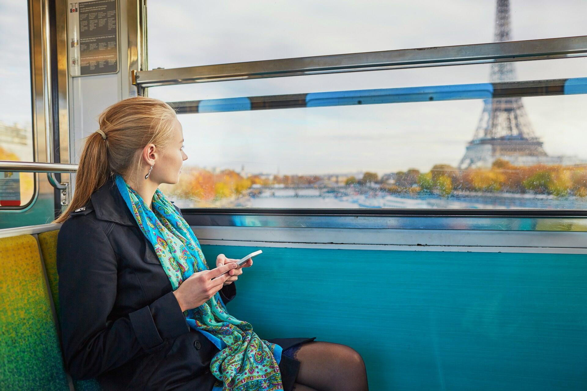 A woman sits on a vibrant train, holding a smartphone while gazing out at the Eiffel Tower and Seine River through the window.