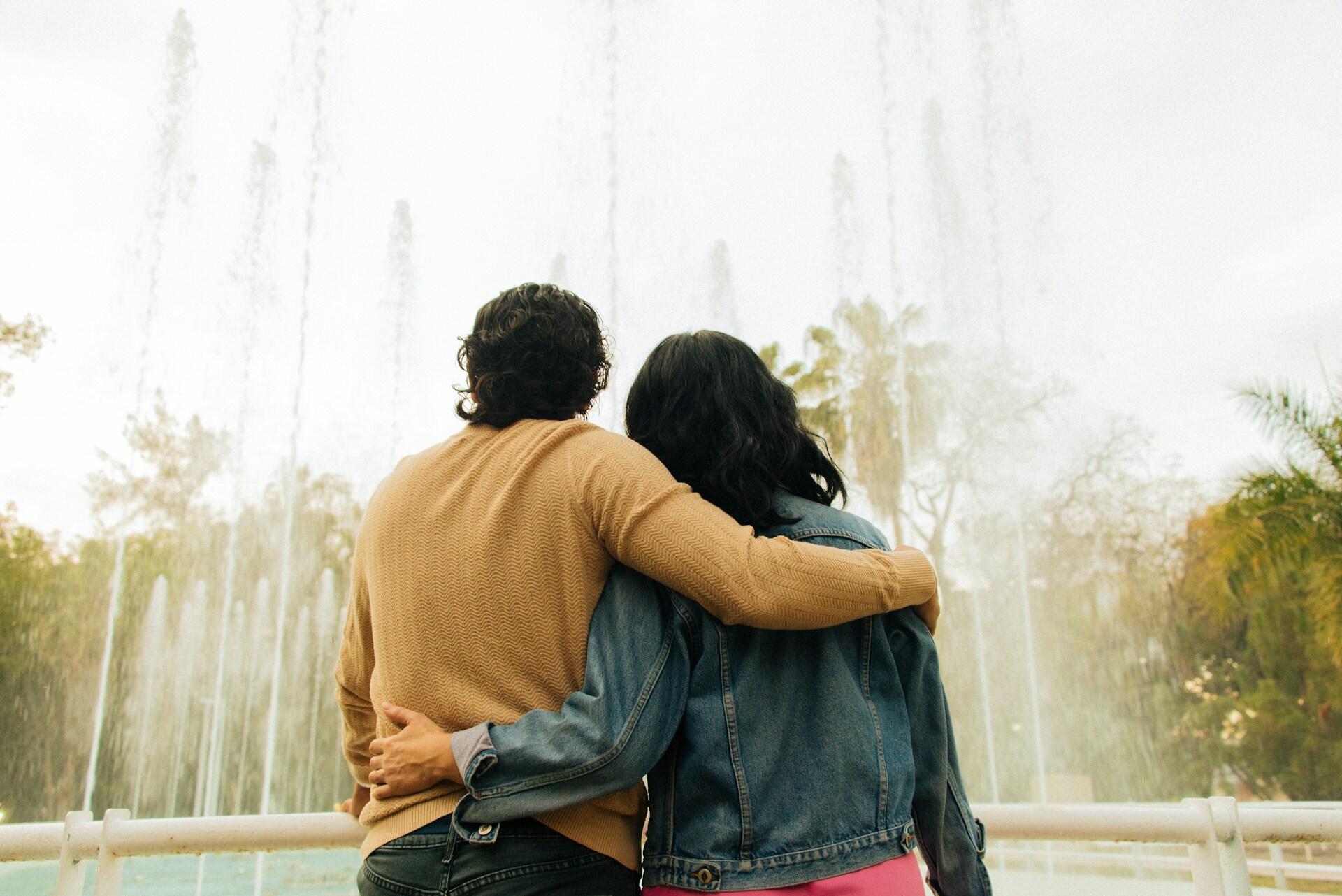 A couple stands together, embracing affectionately, in front of a colourful fountain, surrounded by lush greenery.