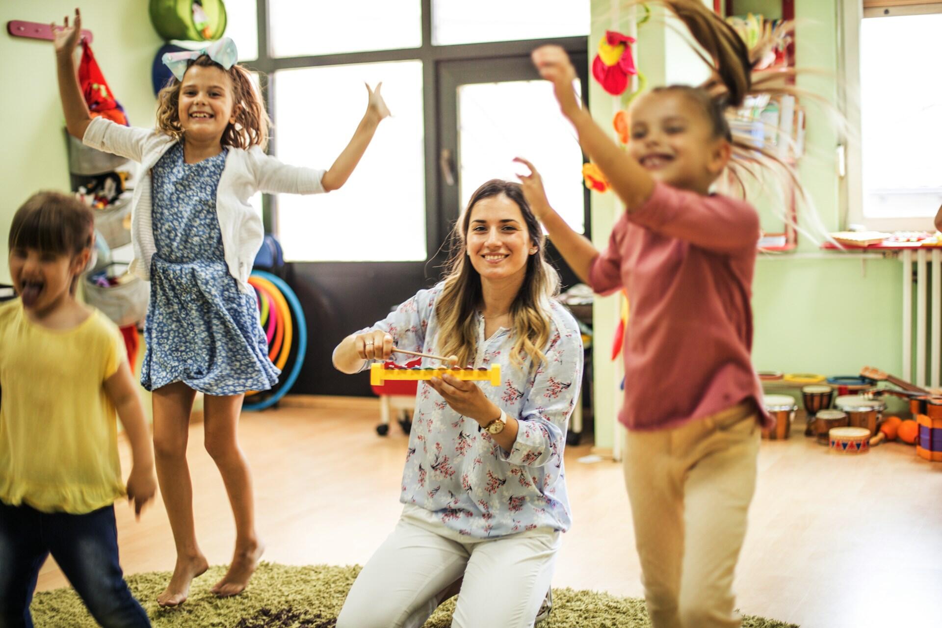 School children dance in class as the teacher kneels on the floor among them.