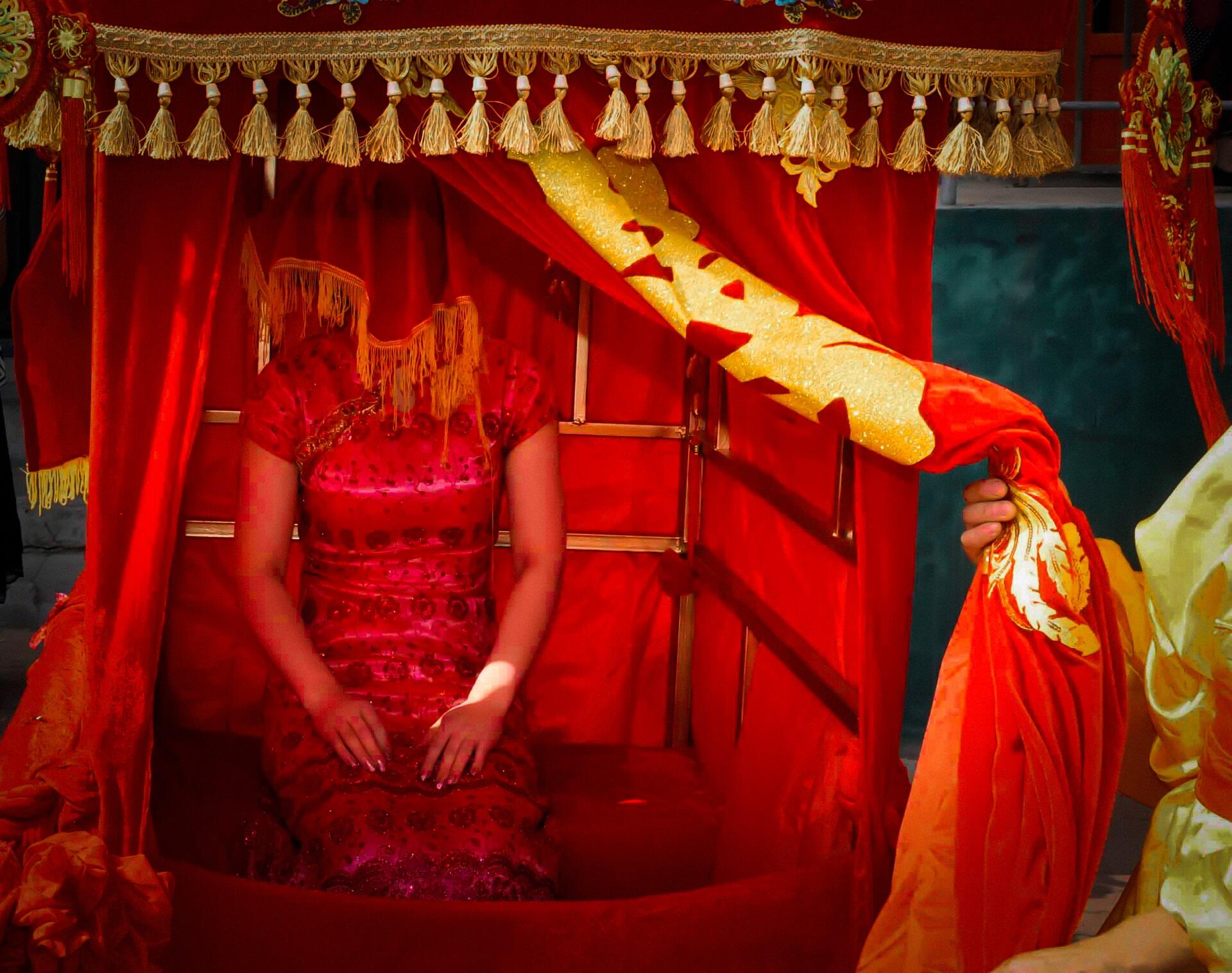 A woman in red with a red veil on her head seated in a red and gold enclosure.