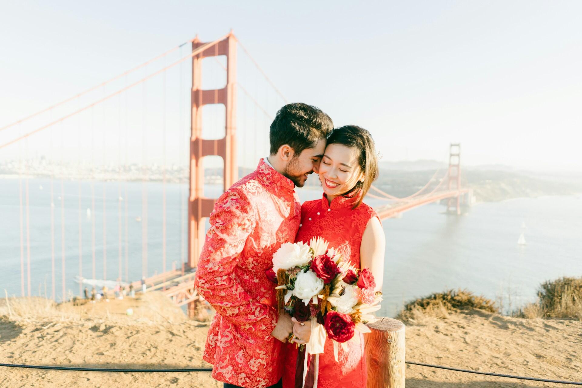 A couple in red traditional Chinese weeding outfits shares a romantic moment, with the Golden Gate Bridge and bay in the background.