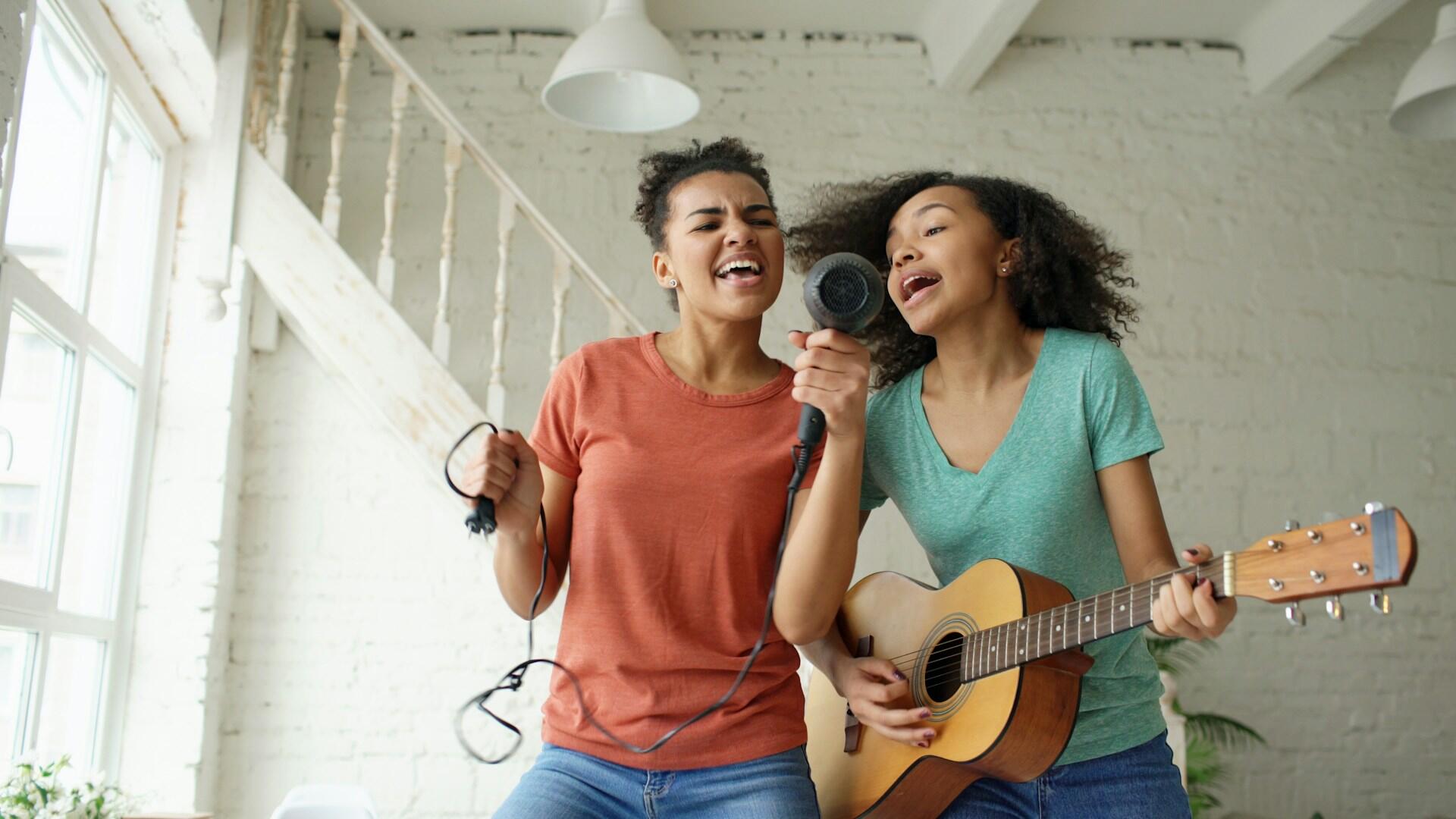 two teen girls playing and pretend singing into a hair dryer while playing guitar