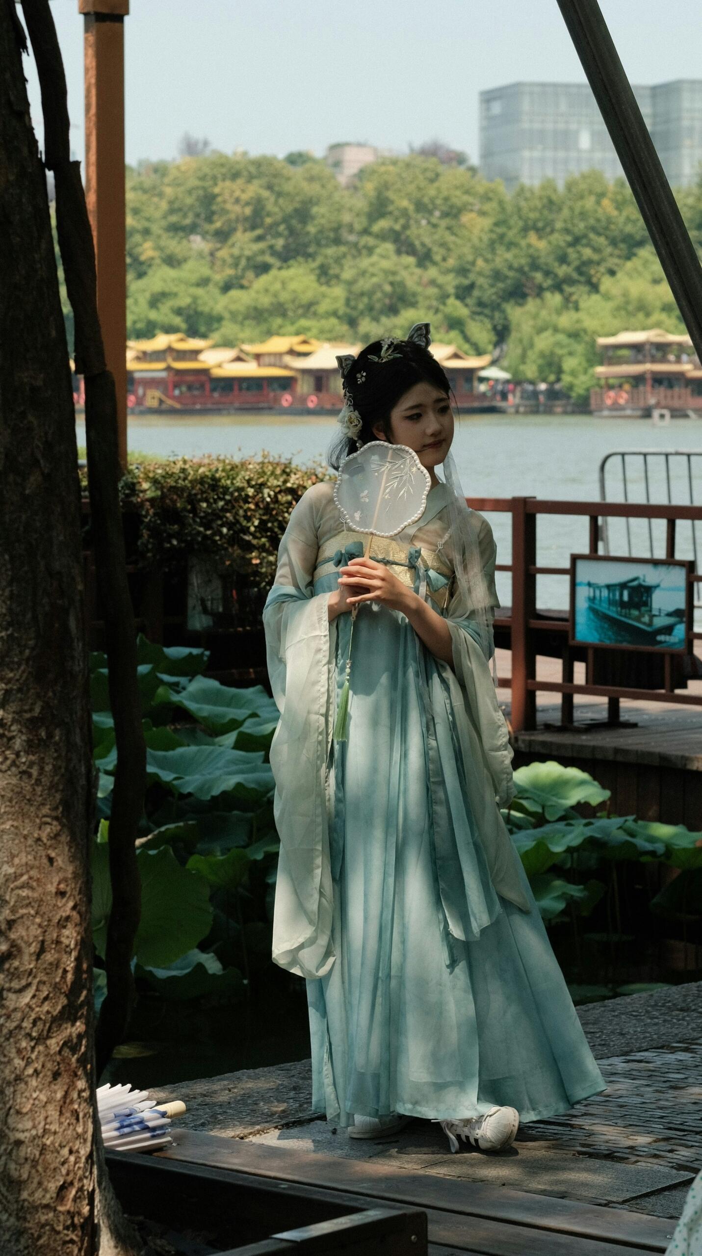 A woman wears elegant robes and holds a fan by a lake.