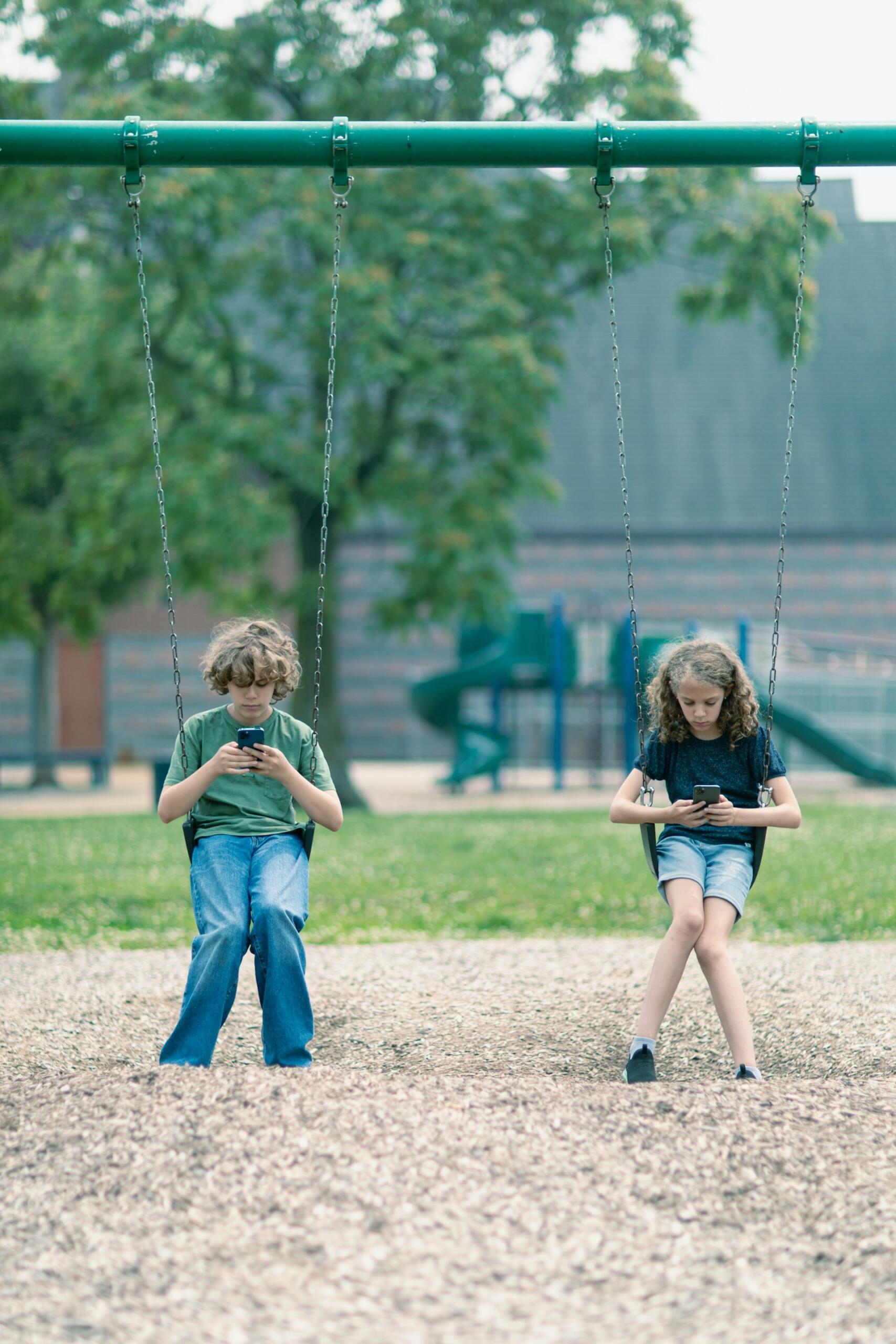 two kids sitting on a swing set using their phones instead of playing