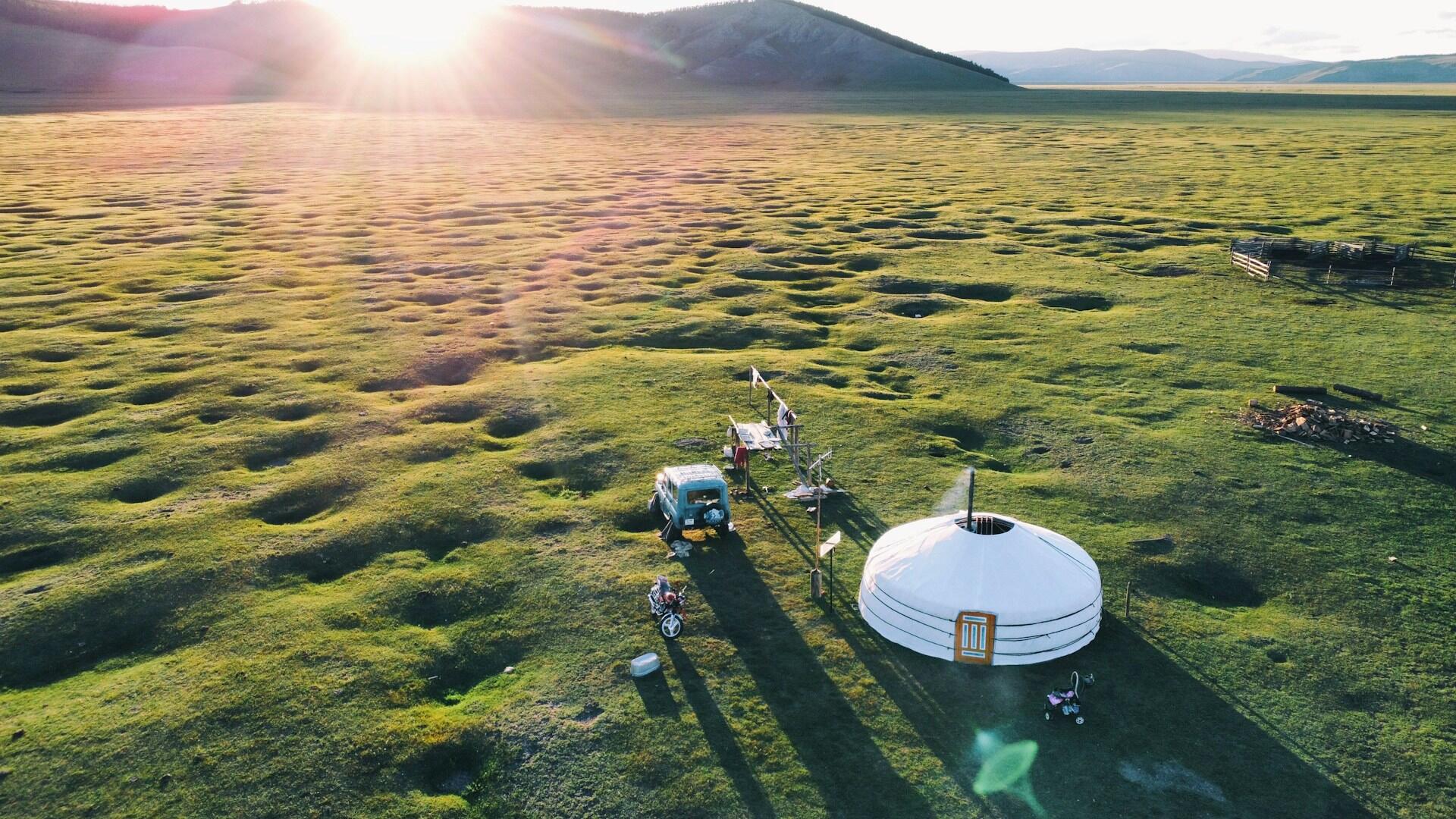A vast expanse of green lawn with a white yurt on it and two vehicles parked in front. The day is sunny and the mood is tranquil.