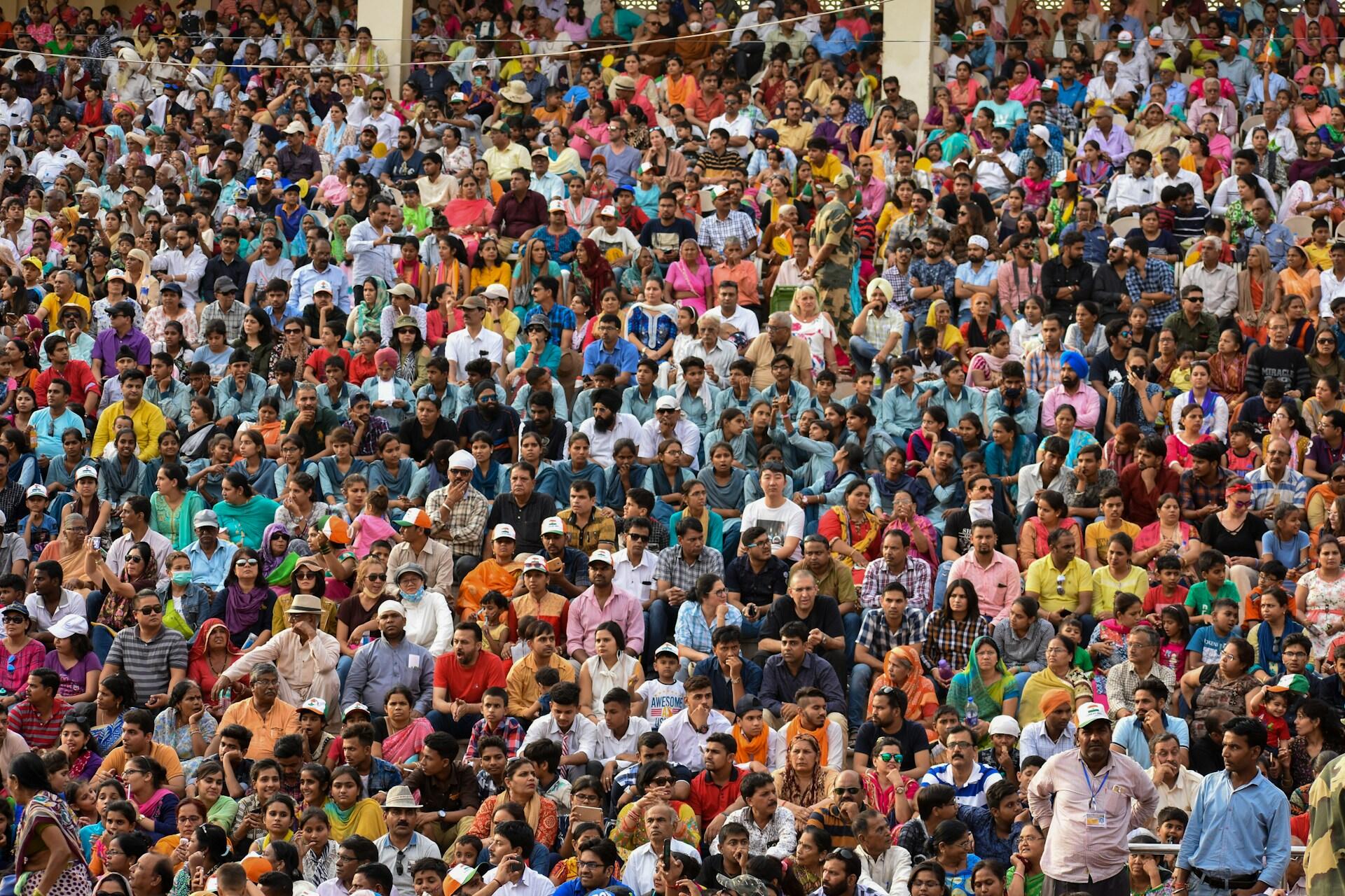 A large crowd wearing many different bright colours gather outdoors. The mood is festive and expectant.