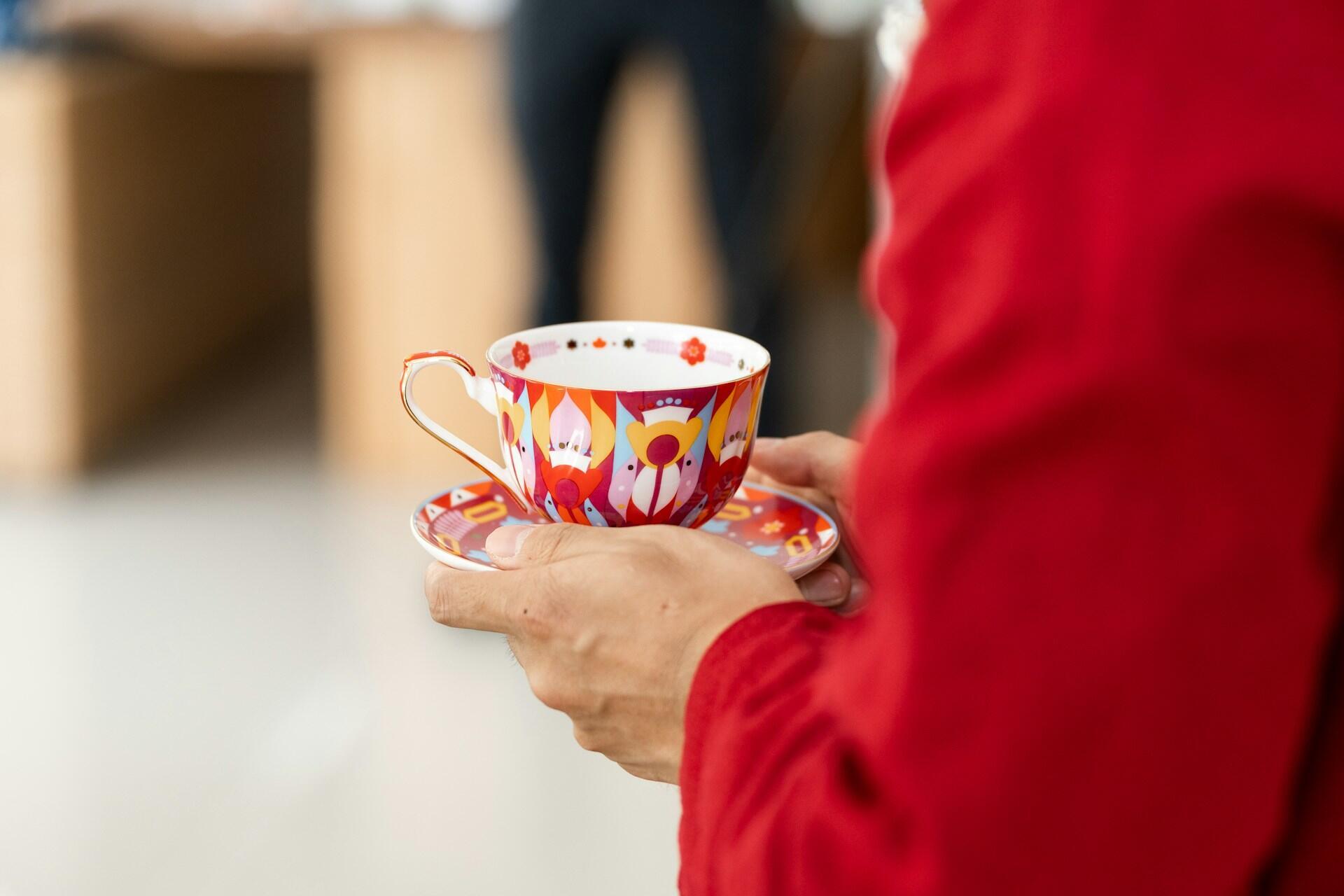 A person wearing red holds a painted teacup on a saucer. 