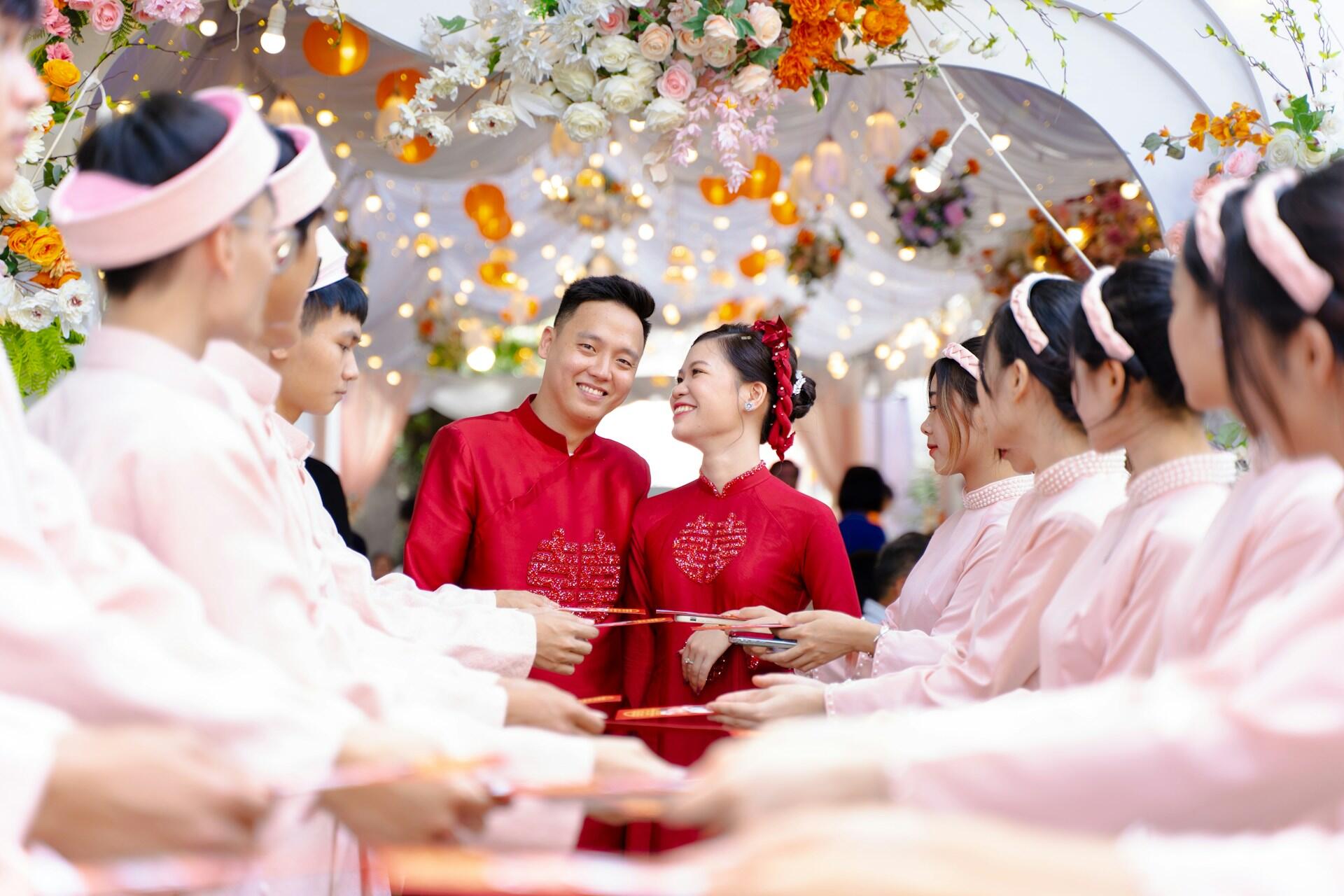A wedding ceremony with a vibrant floral backdrop, featuring couples in traditional wedding attire surrounded by attendants holding red envelopes.
