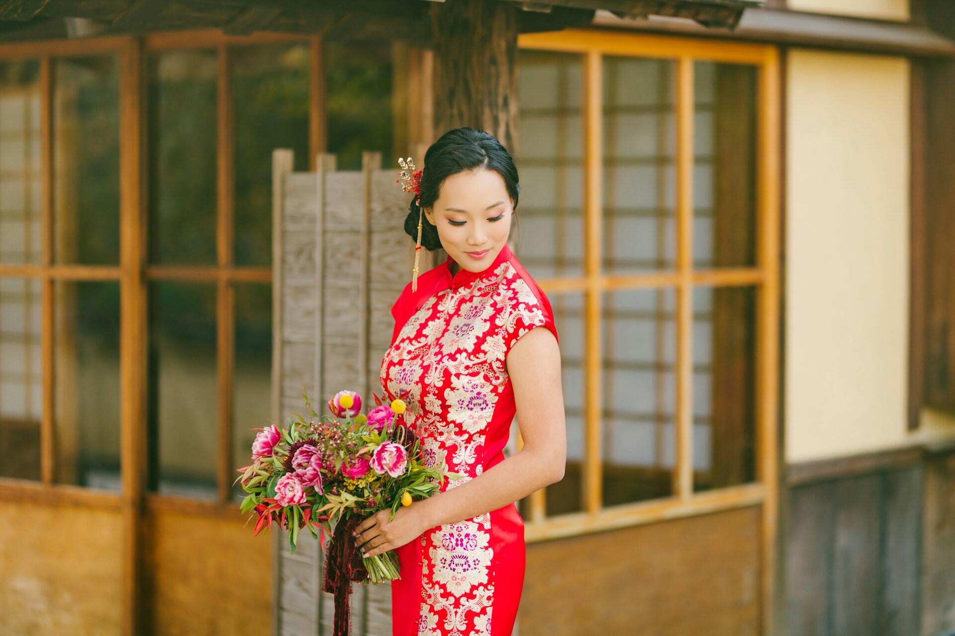 A woman in a traditional Chinese red qipao adorned with intricate patterns holds a bouquet of flowers.
