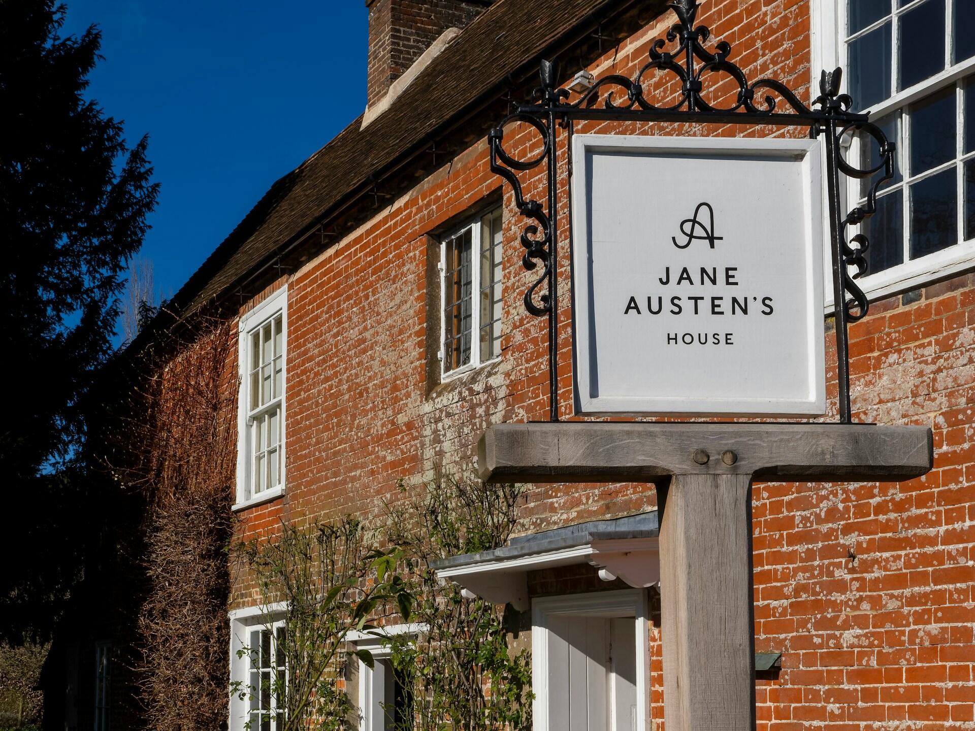 Sign for Jane Austen's House, featuring a white background and elegant lettering, set against a rustic brick home.
