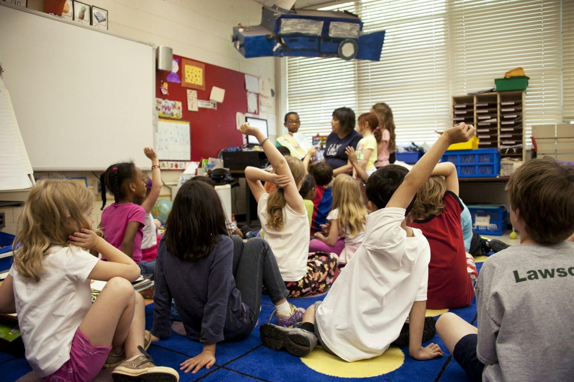 Children sitting on a classroom floor raising their hands while a teacher speaks.