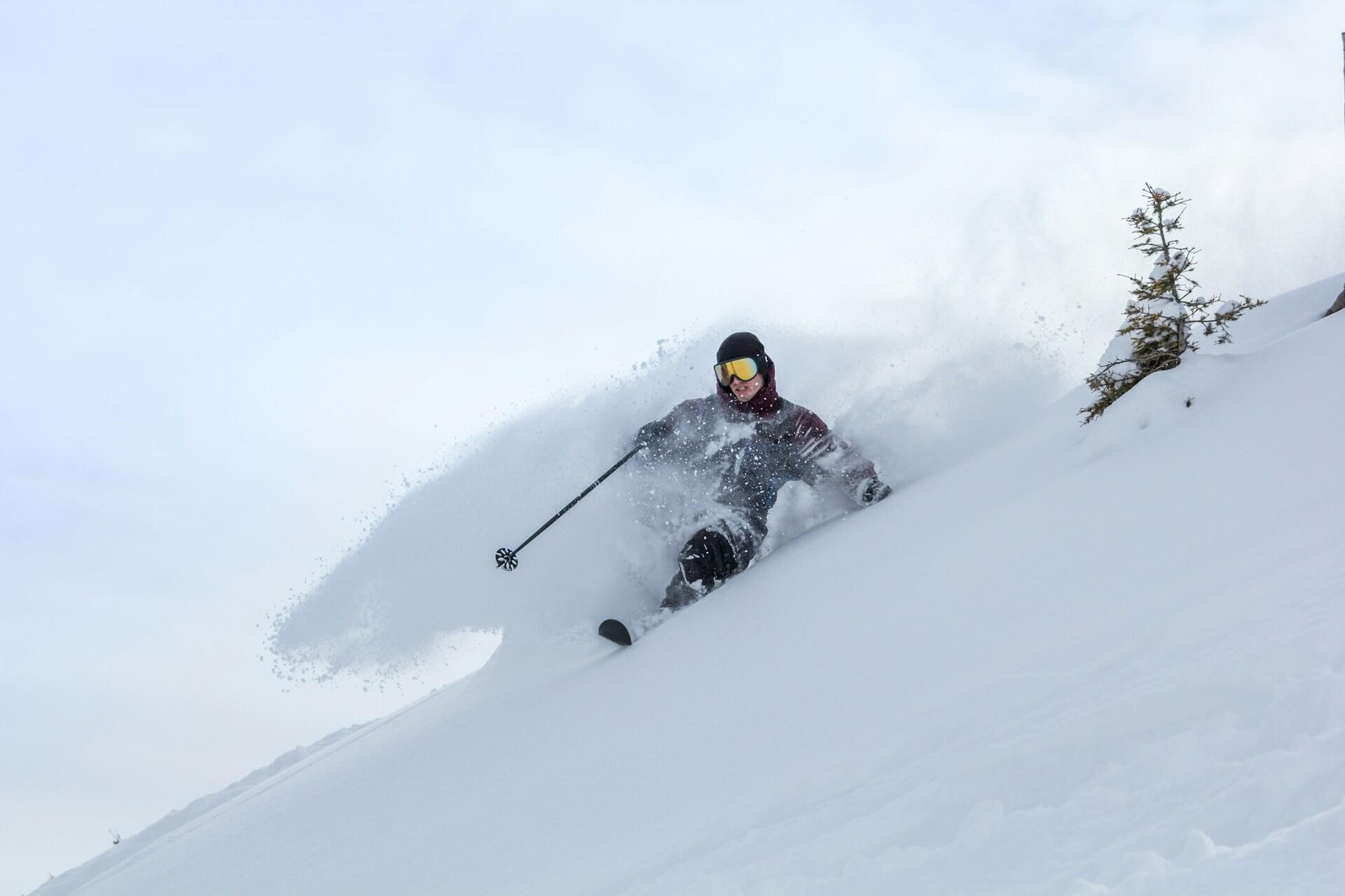 A skier expertly maneuvers through deep powder snow, sending up a spray of snow against a cloudy sky.