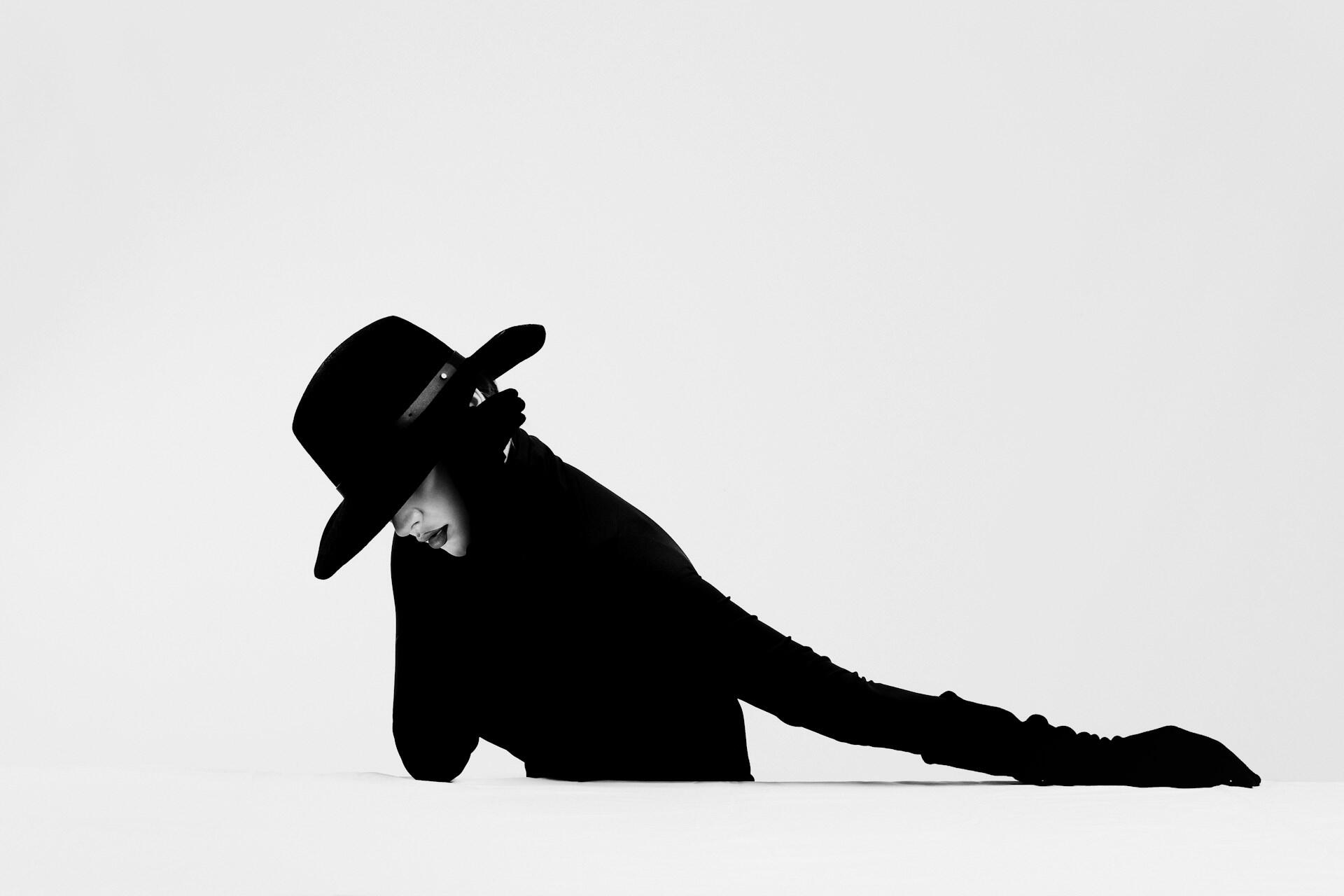 A fashion model draped in a black hat and clothing leans over a white surface.