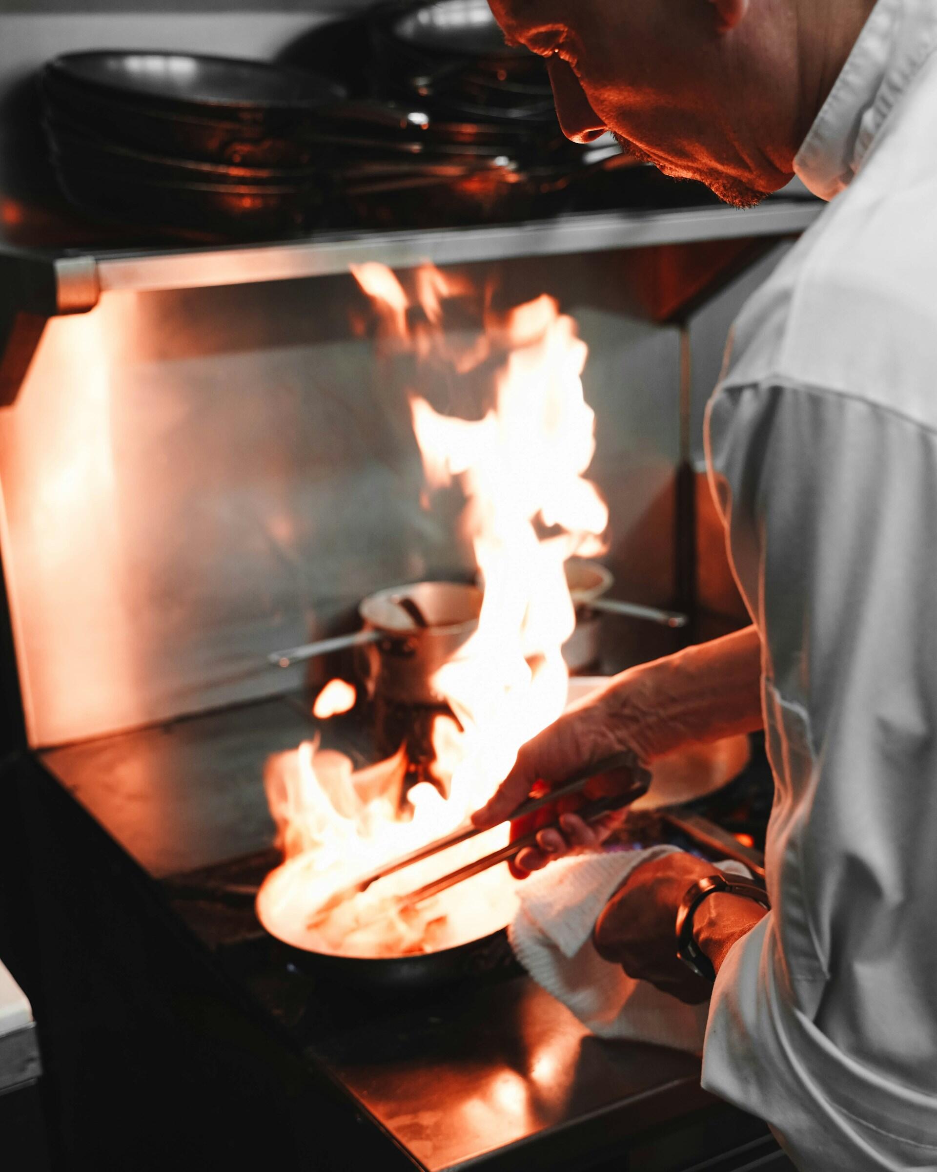 A chef in white jacket cooking with flames rising from a pan on a professional stove.