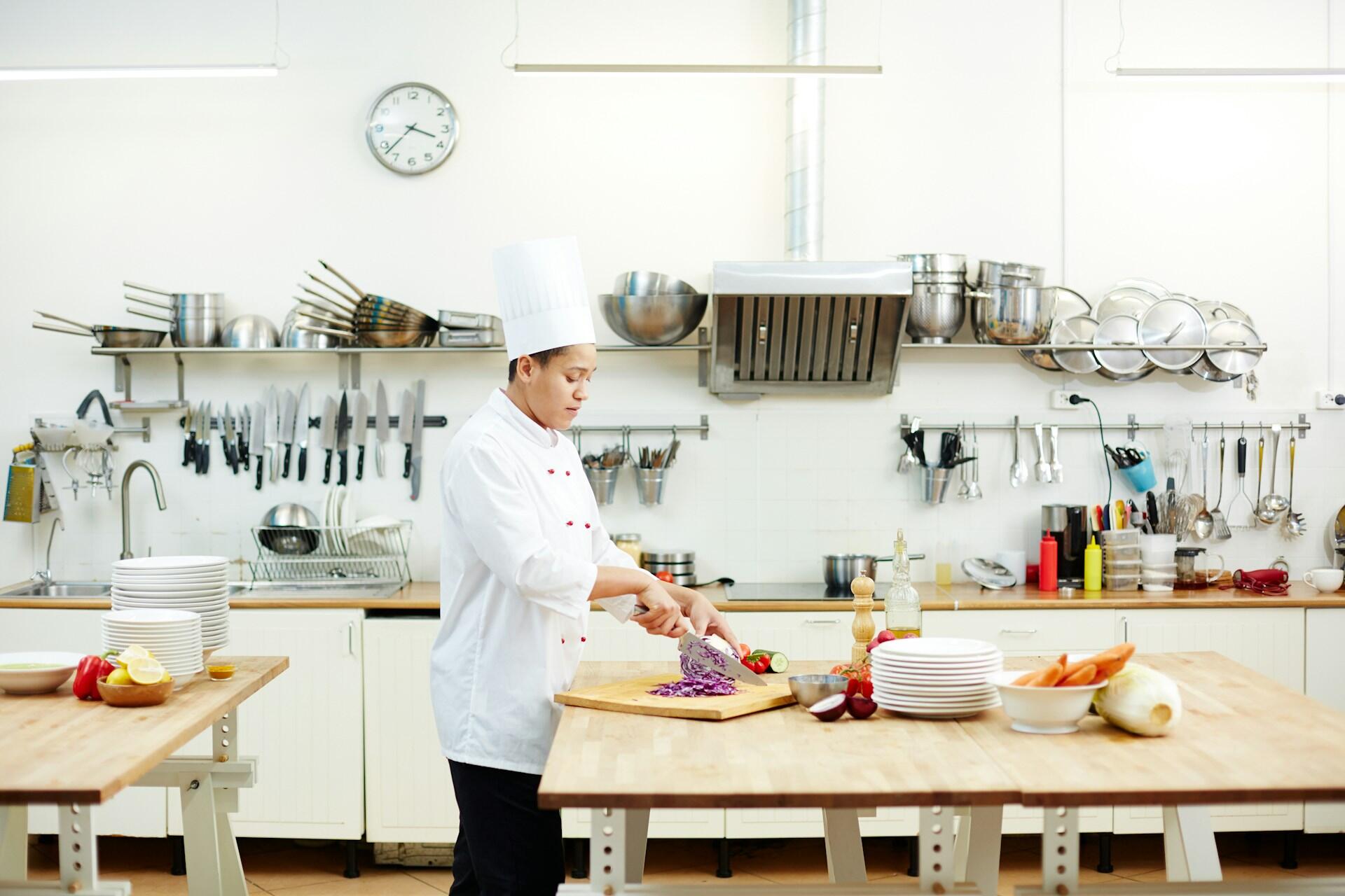 A cook in white uniform chopping purple cabbage on a wooden cutting board in a professional kitchen.