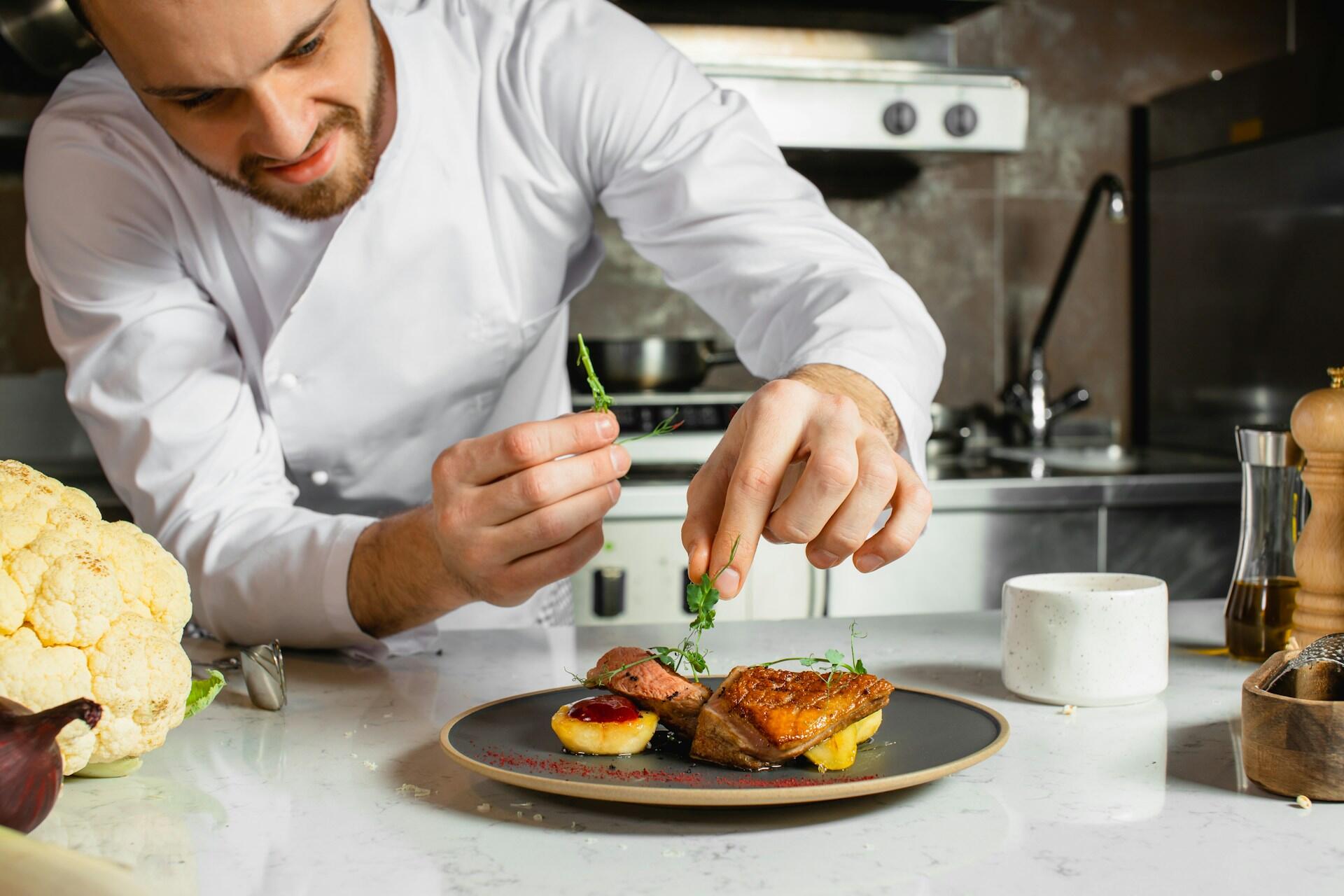 A chef garnishes a beautifully plated dish featuring meat and sauce.