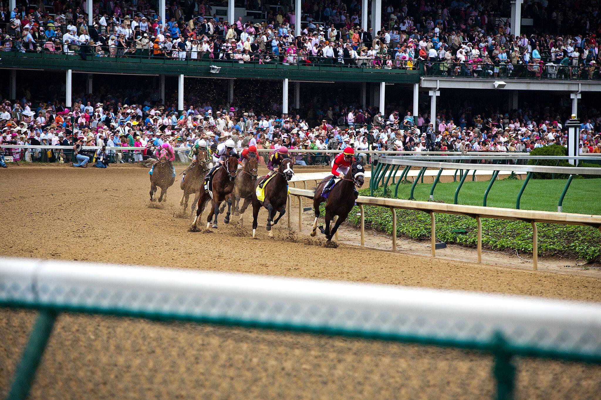 A horse race in progress, with jockeys and horses competing on a sandy track, surrounded by a large crowd in the stands.