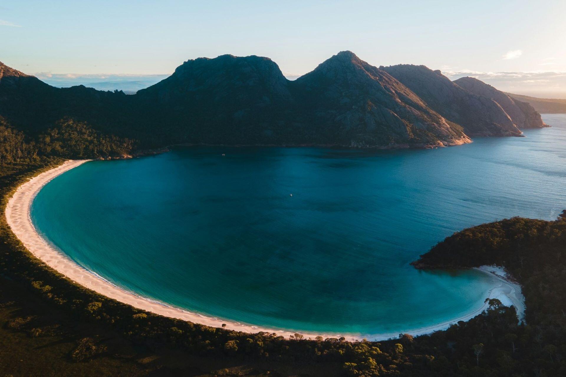 Aerial view of a curved sandy beach and turquoise bay surrounded by mountains in Australia