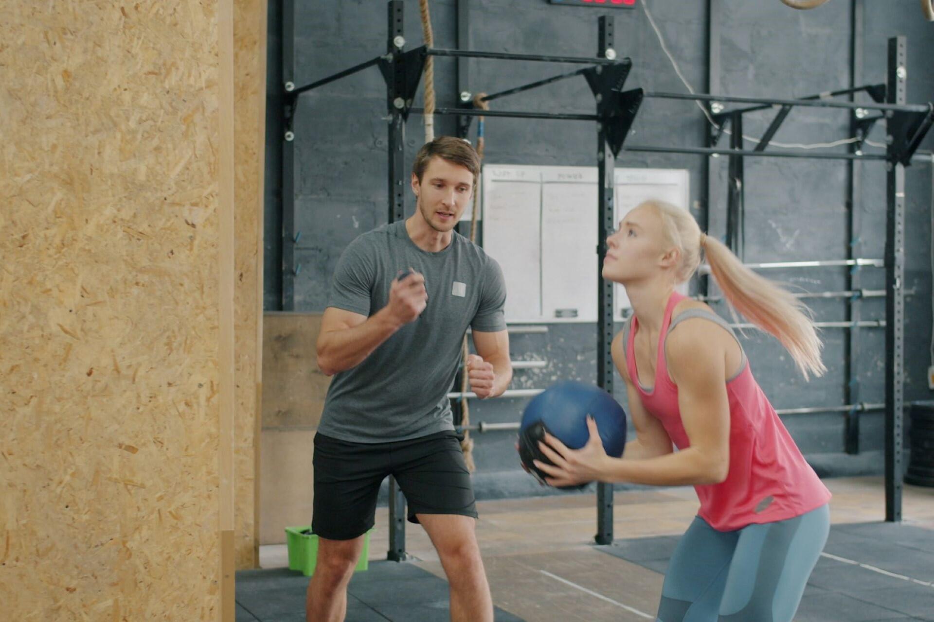 Athlete performing wall ball exercise in a functional fitness gym while a coach observes during hybrid training