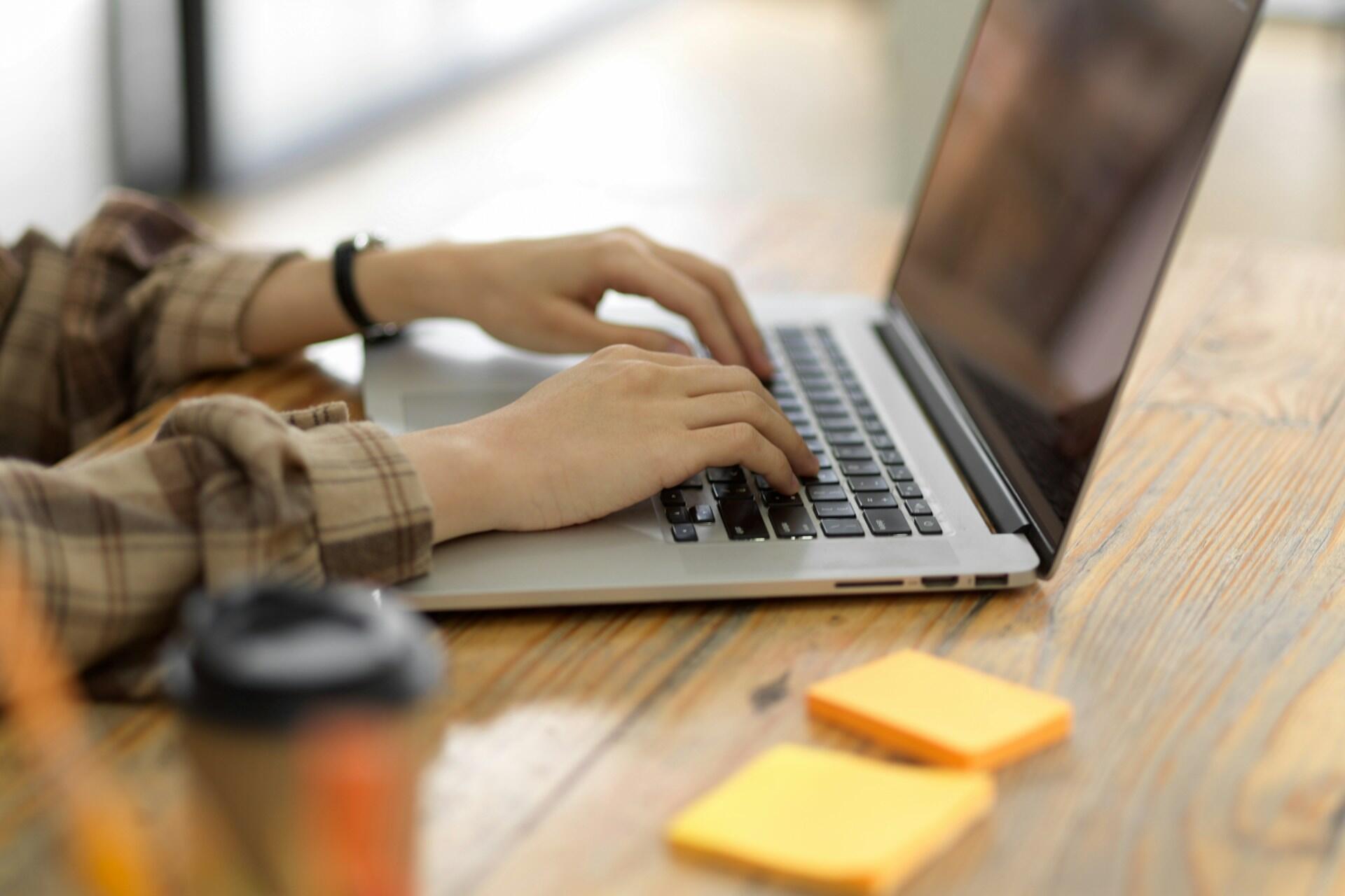 Hands typing on a laptop keyboard with a wooden table, two yellow sticky notes, and a coffee cup.