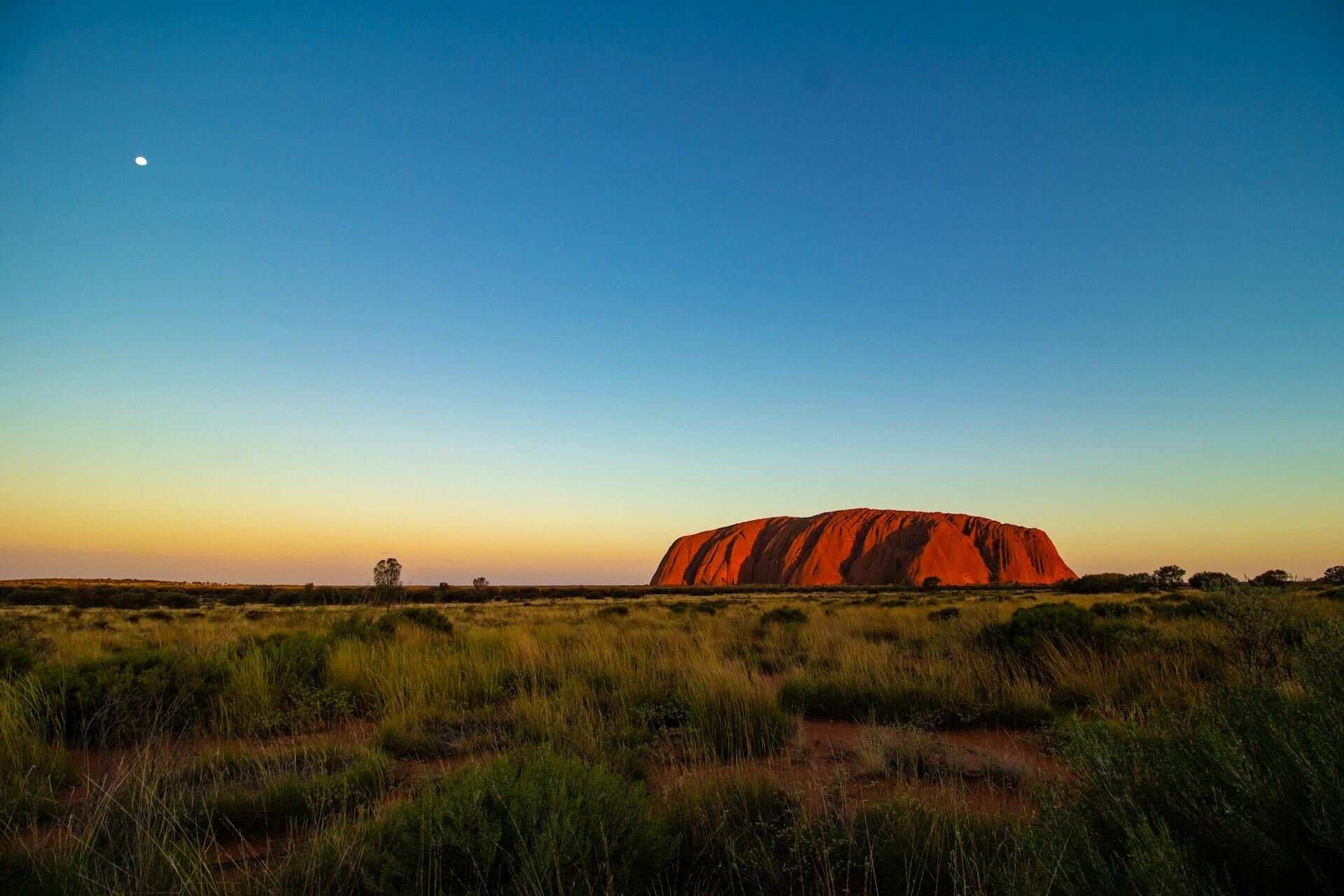 Uluru rock formation in the Australian outback with grassland and sunset sky