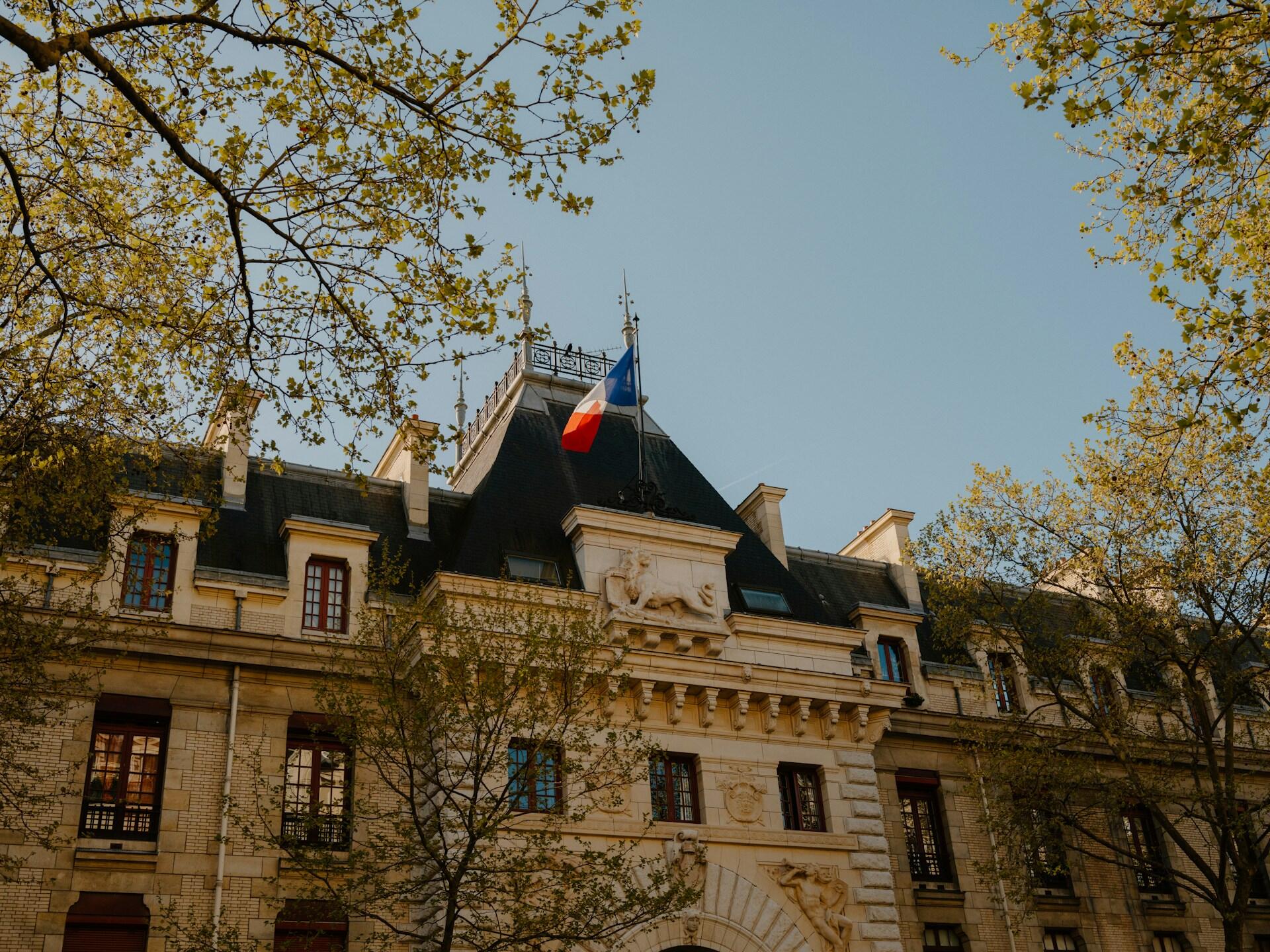 Historic stone building with a French flag on the roof, surrounded by trees under a clear blue sky.