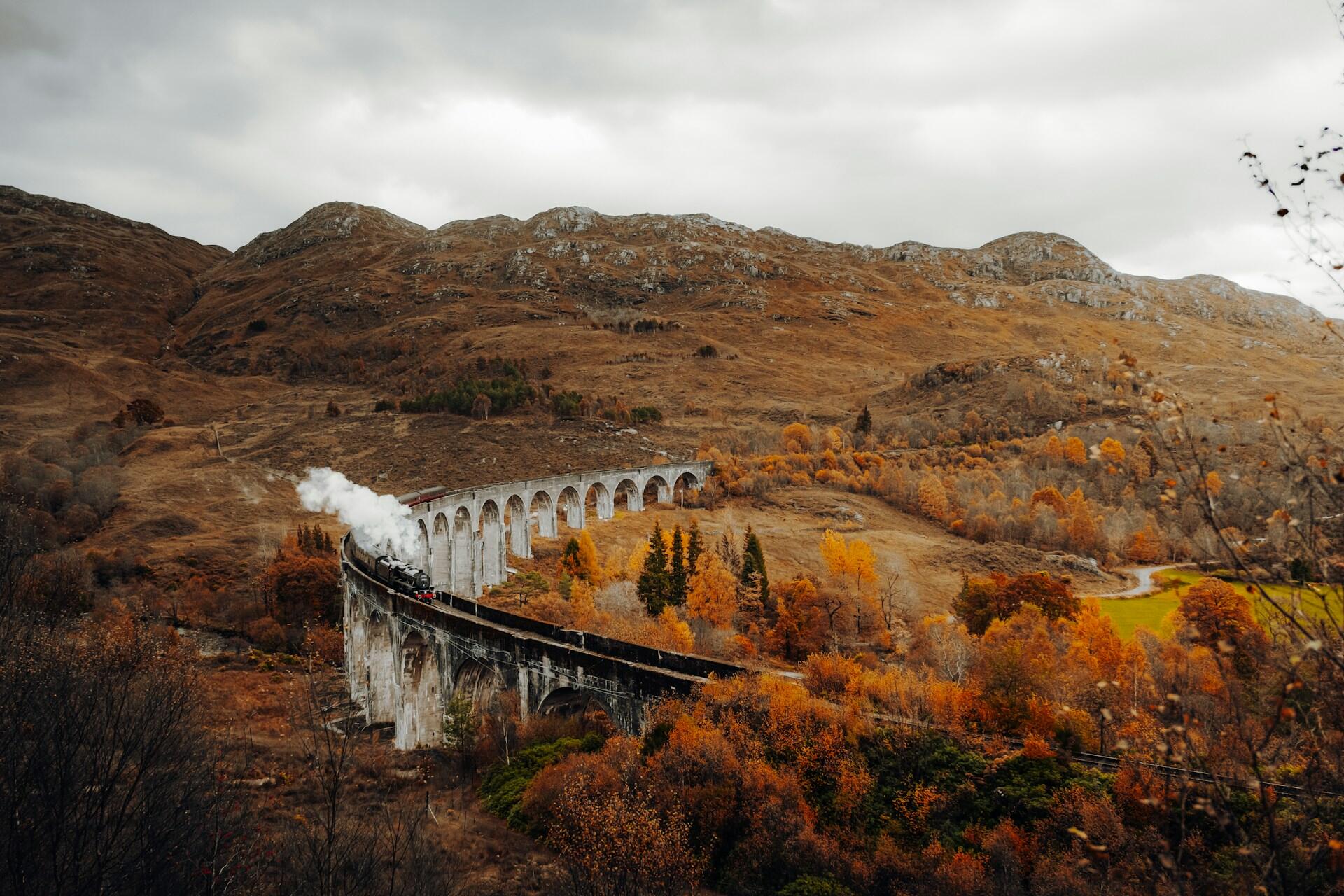 Steam train crossing a curved stone viaduct in autumnal mountainous landscape.
