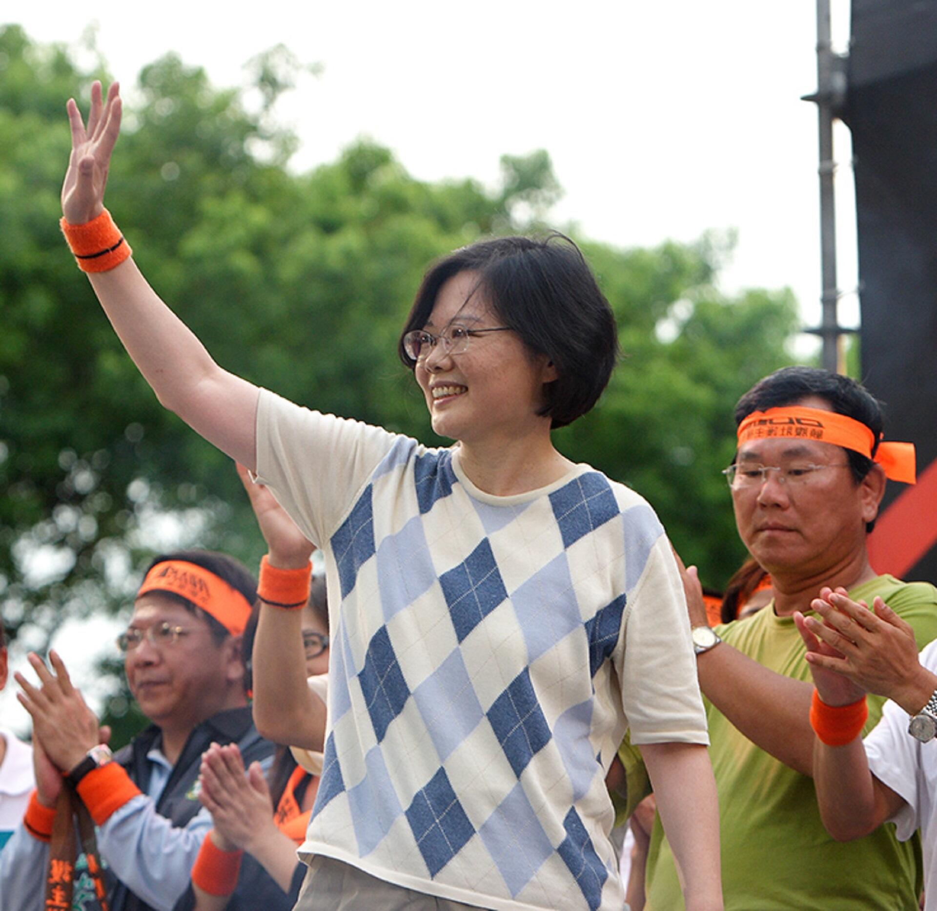 A woman with short black hair, wearing glasses and a blue and tan jumper, waves at people in the distance.