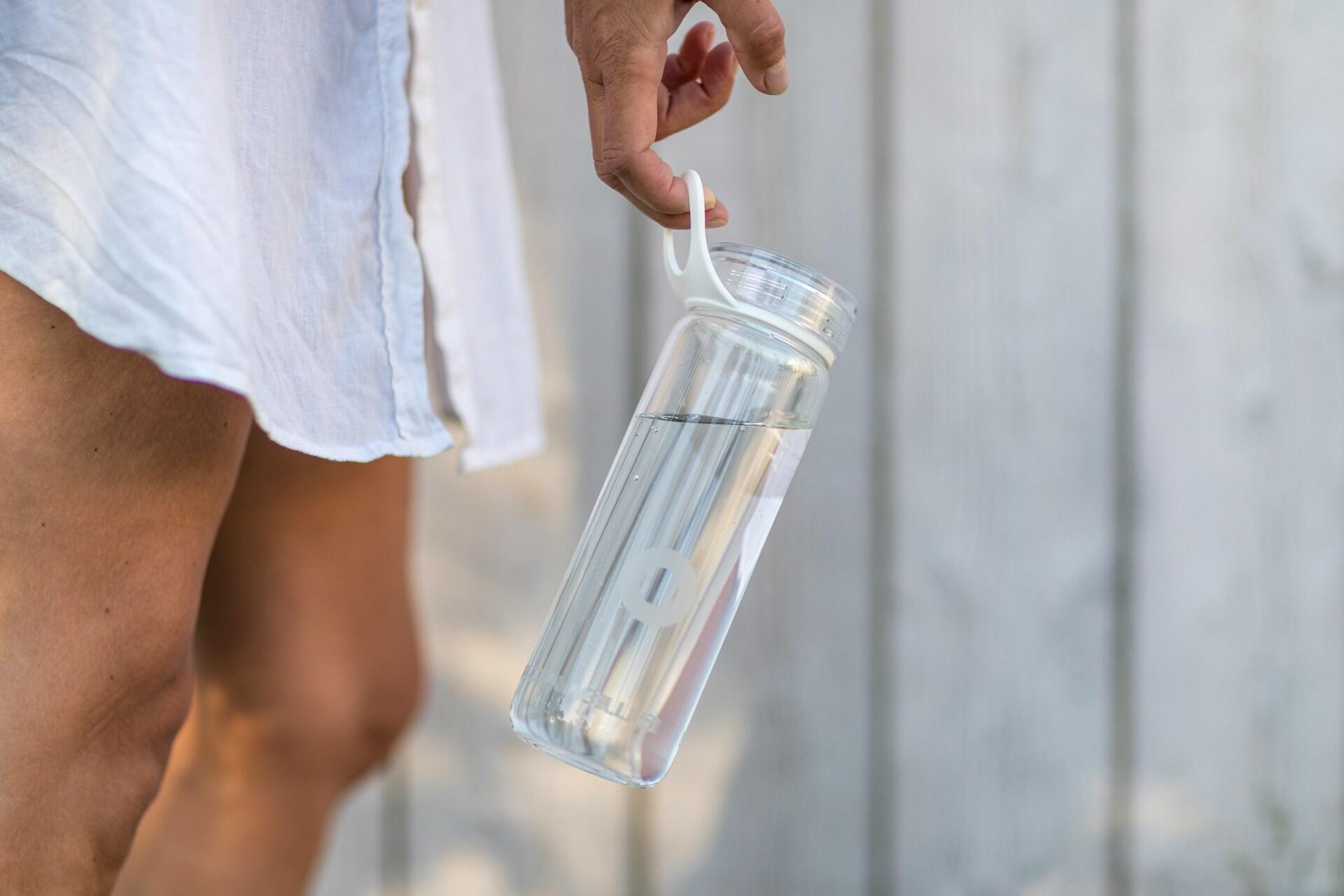 A woman wearing a white shirt holds a clear water bottle by its carrying strap.