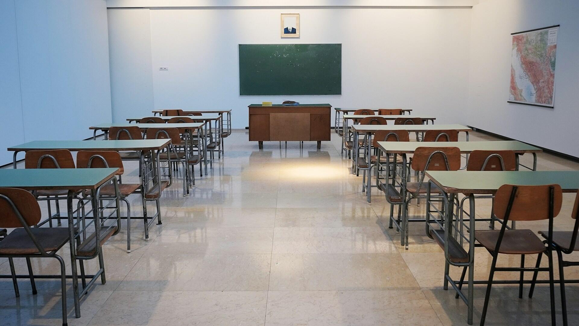 An empty classroom with two-place desks on either side of the room, separated by a wide aisle. A teacher's desk sits at the front of the room, under a chalkboard.