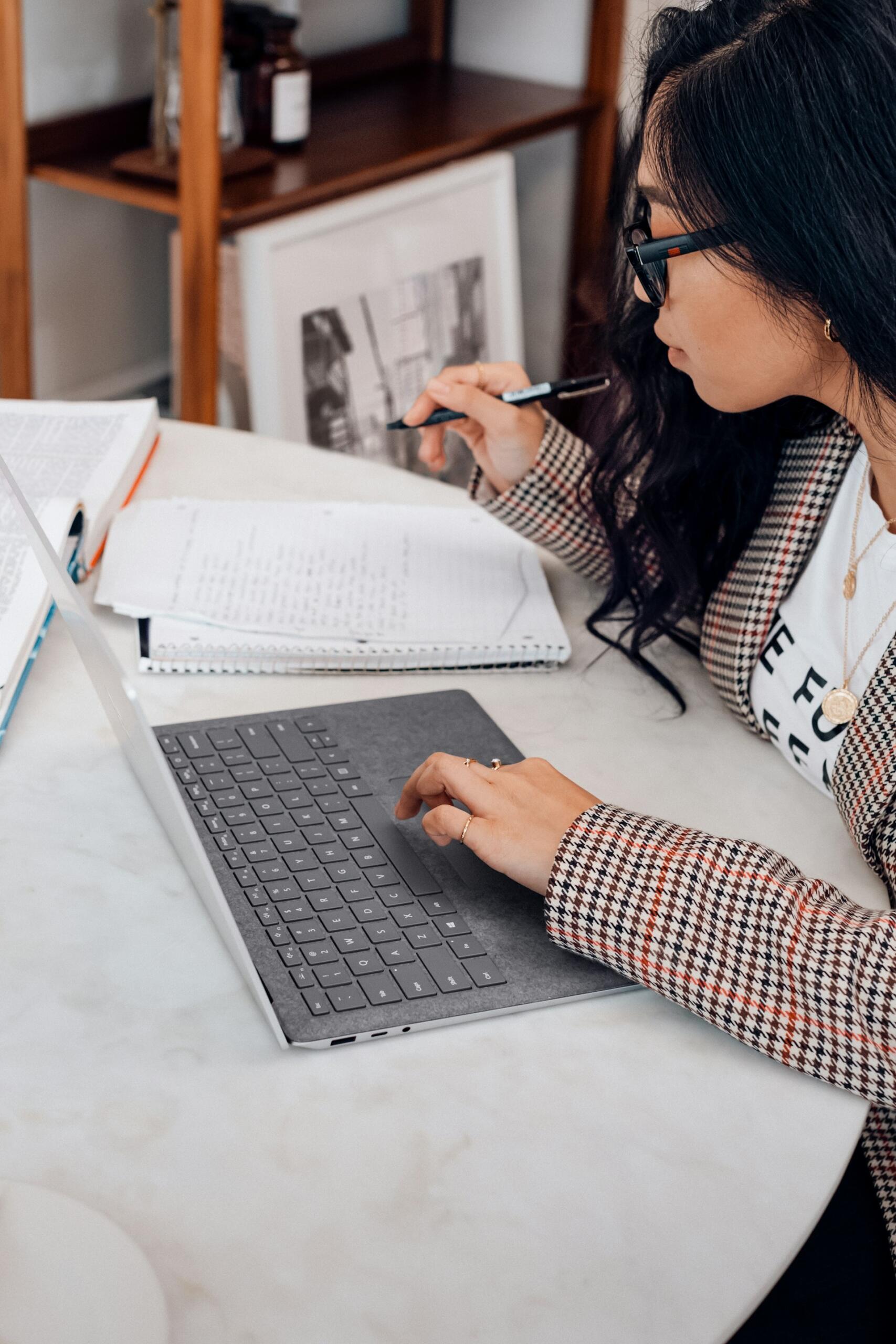 A woman with long black hair, wearing glasses and a long-sleeved jumper, holds a pen in her hand while working a computer with her other hand.