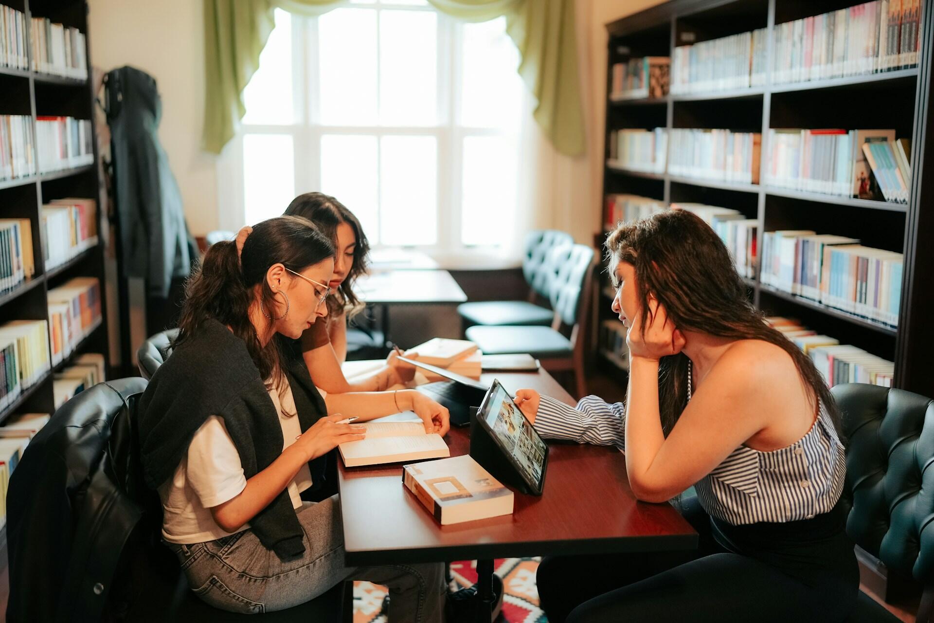 Three young women in a library with study materials open in front of them, and one holding a tablet up to read from.