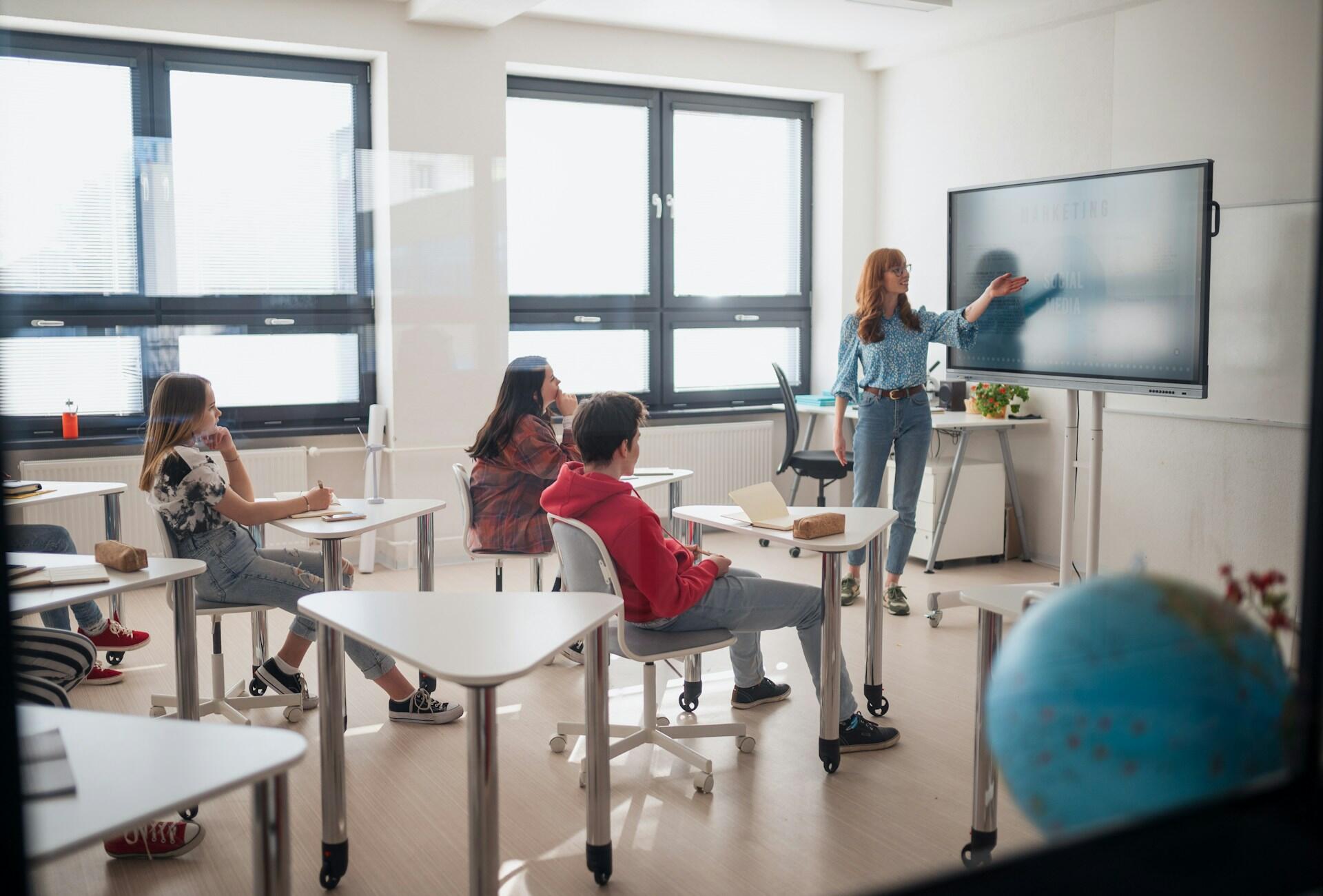 A woman wearing jeans and a blue shirt stands at the head of the classroom, pointing at the board, while students sit at white triangular desks.
