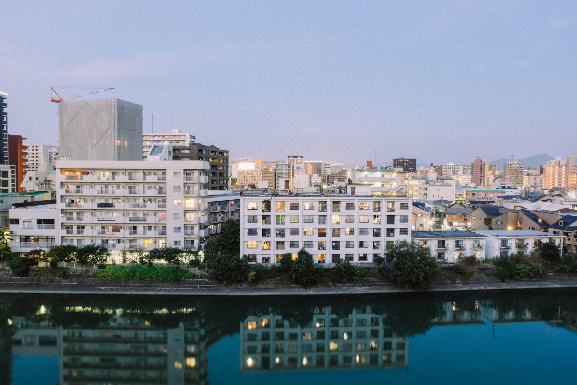 A collection of residential buildings painted white fronting a calm river, with greenery on its banks.
