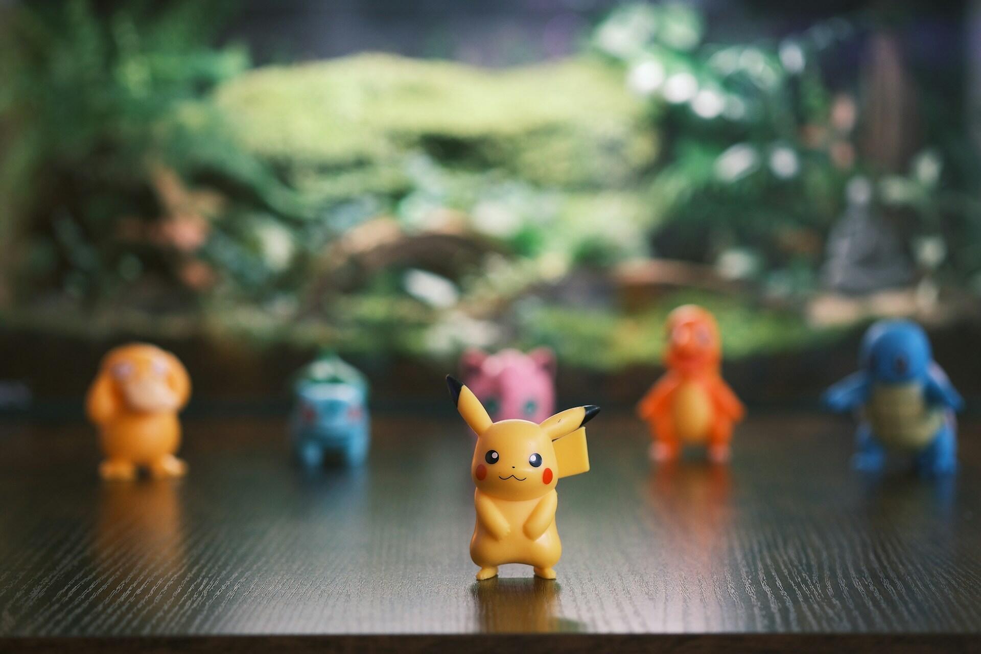 Small Pikachu toy figure stands in focus on a wooden surface, with blurry Pokémon figures in the background.