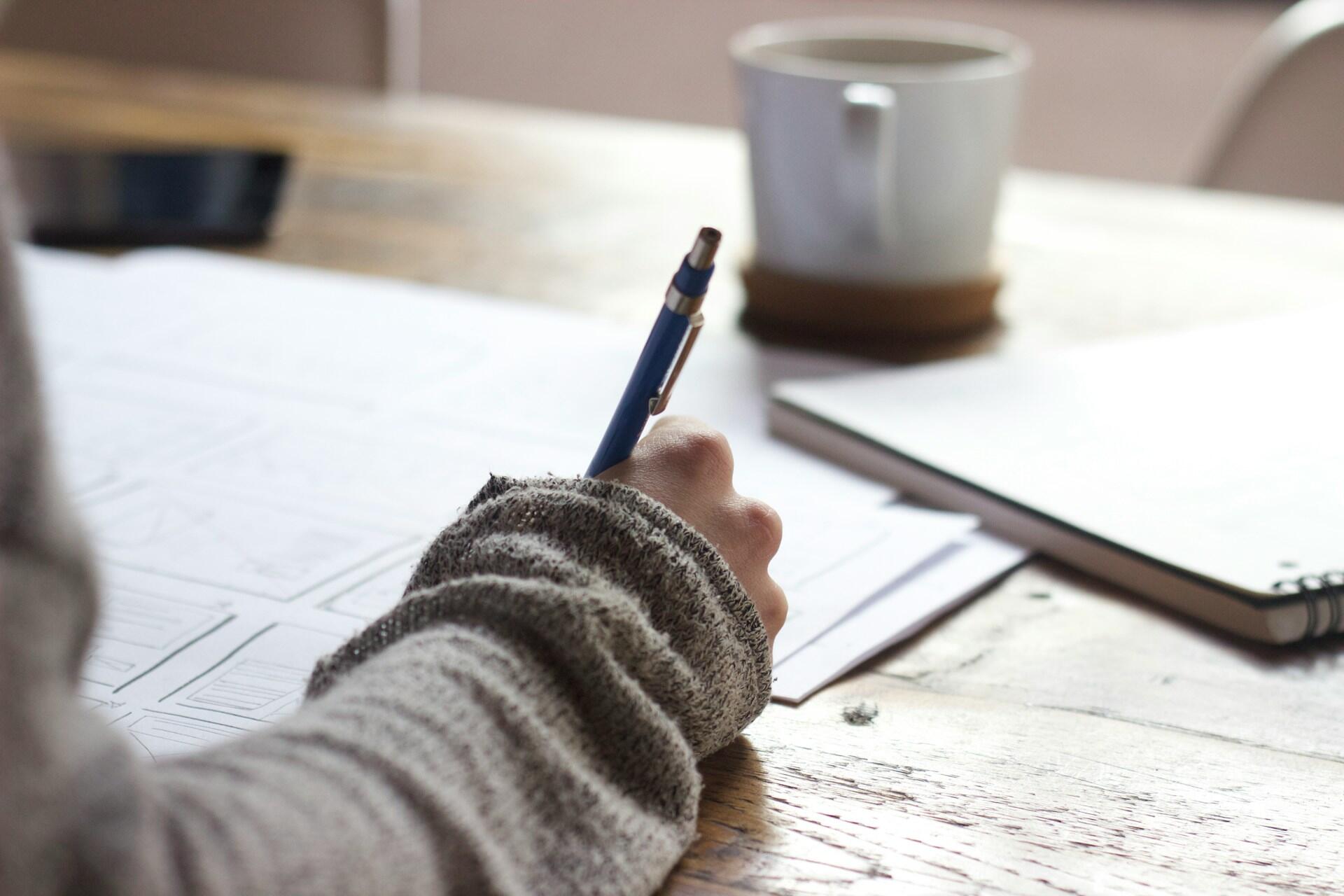 A person wearing a jumper with long sleeves holds a pencil and writes in a notepad, with a cup of drink nearby.
