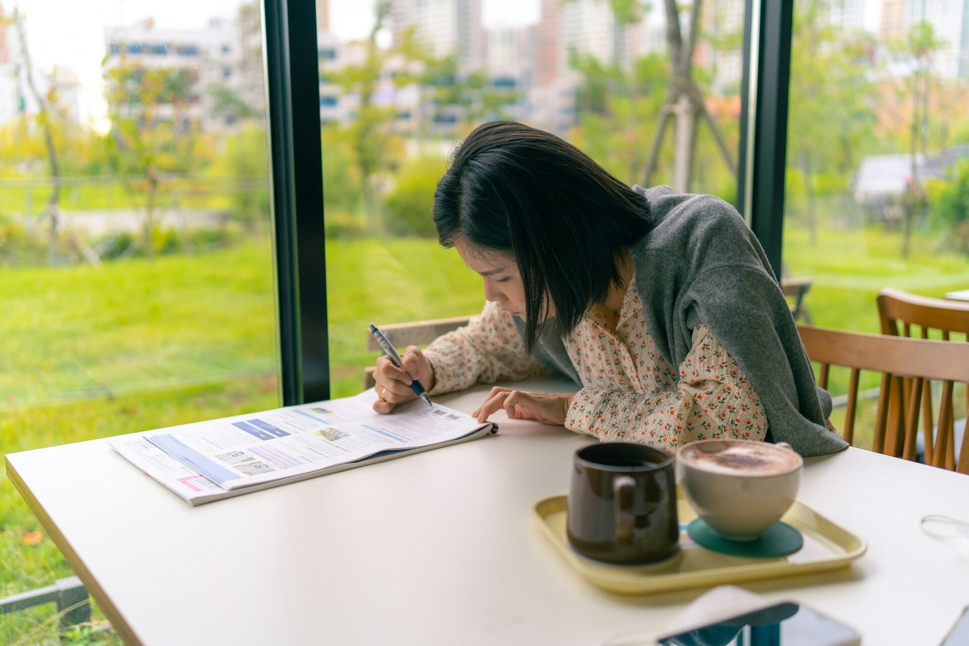 A woman seated at a table on a covered patio, holding a pencil in her right hand and focused on a paper in front of her. She has a coffee by her left arm.