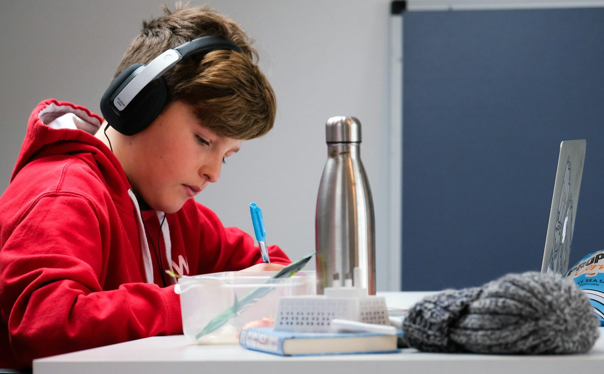 A young man wearing a red hoodies has black headphones on as he writes in a notebook. A steel bottle sits nearby.