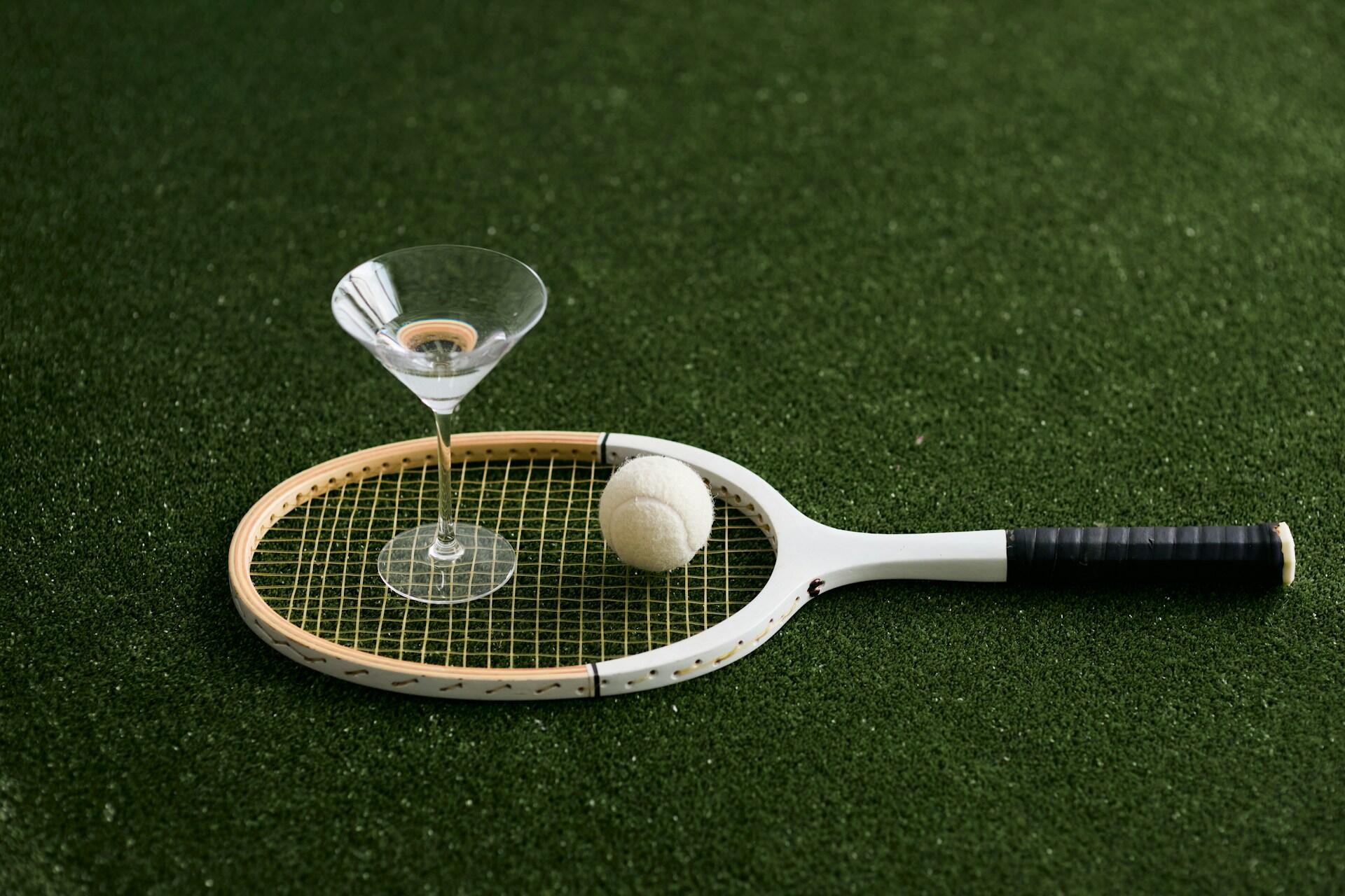 Tennis racket with ball and empty martini glass resting on green grass surface.