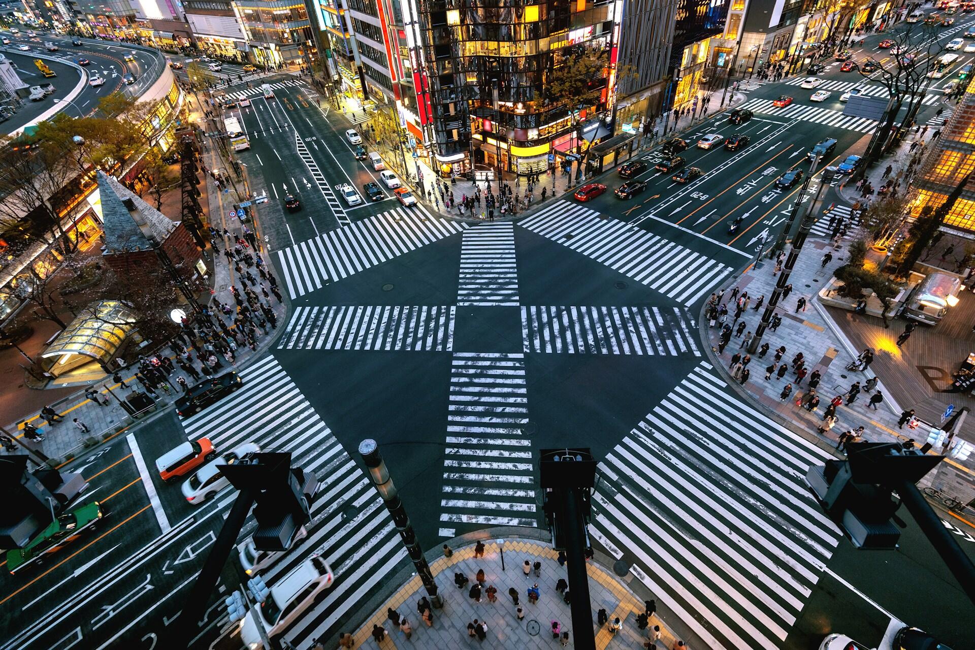 The six-way crosswalk in Tokyo at night, lit up by nearby tall buildings, with a few cars waiting to enter.
