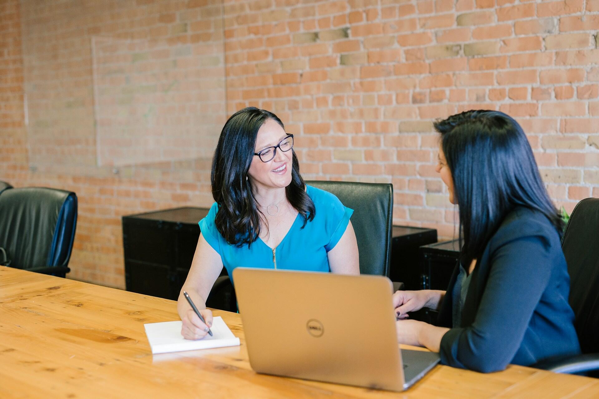 Two women sit at a wooden table with a laptop on it; one woman holds a pen. The mood is friendly and relaxed.