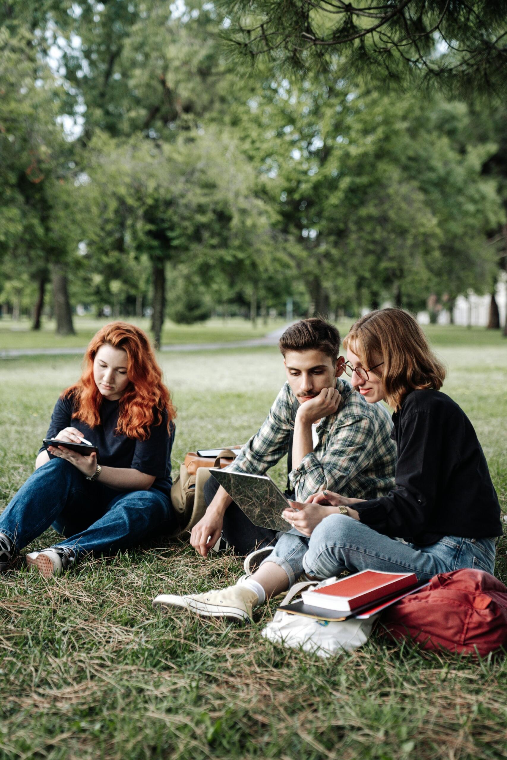Three young people sit on the ground in a park, working on their studies.
