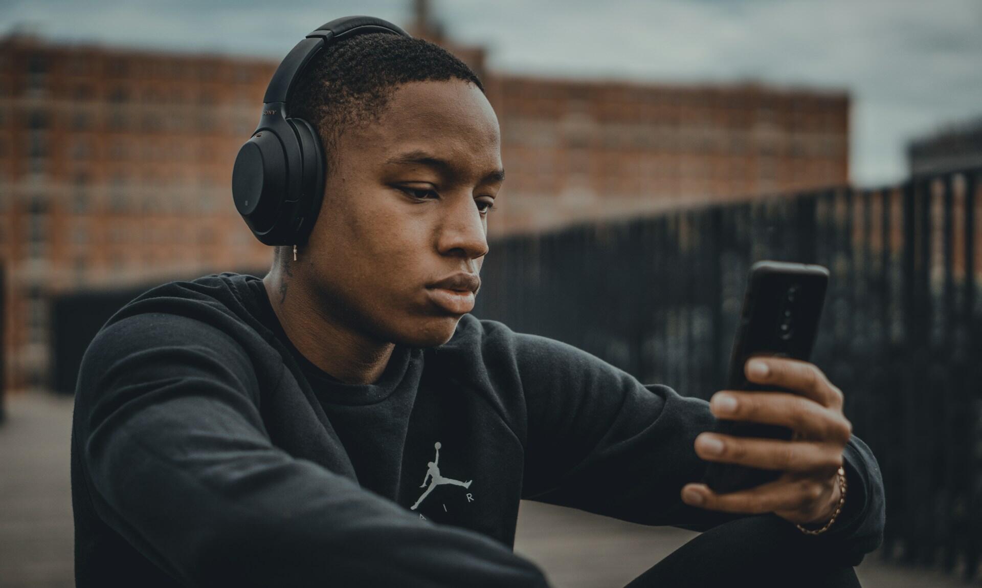 A person wears black headphones while looking at his phone with a serious expression on his face.