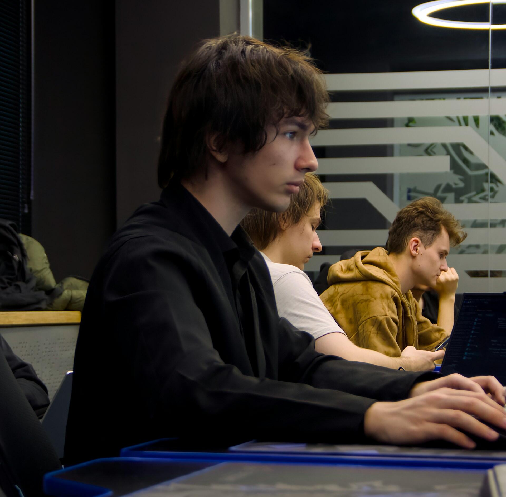 A student wearing a dark top stares intently at a computer screen while other students look bored.