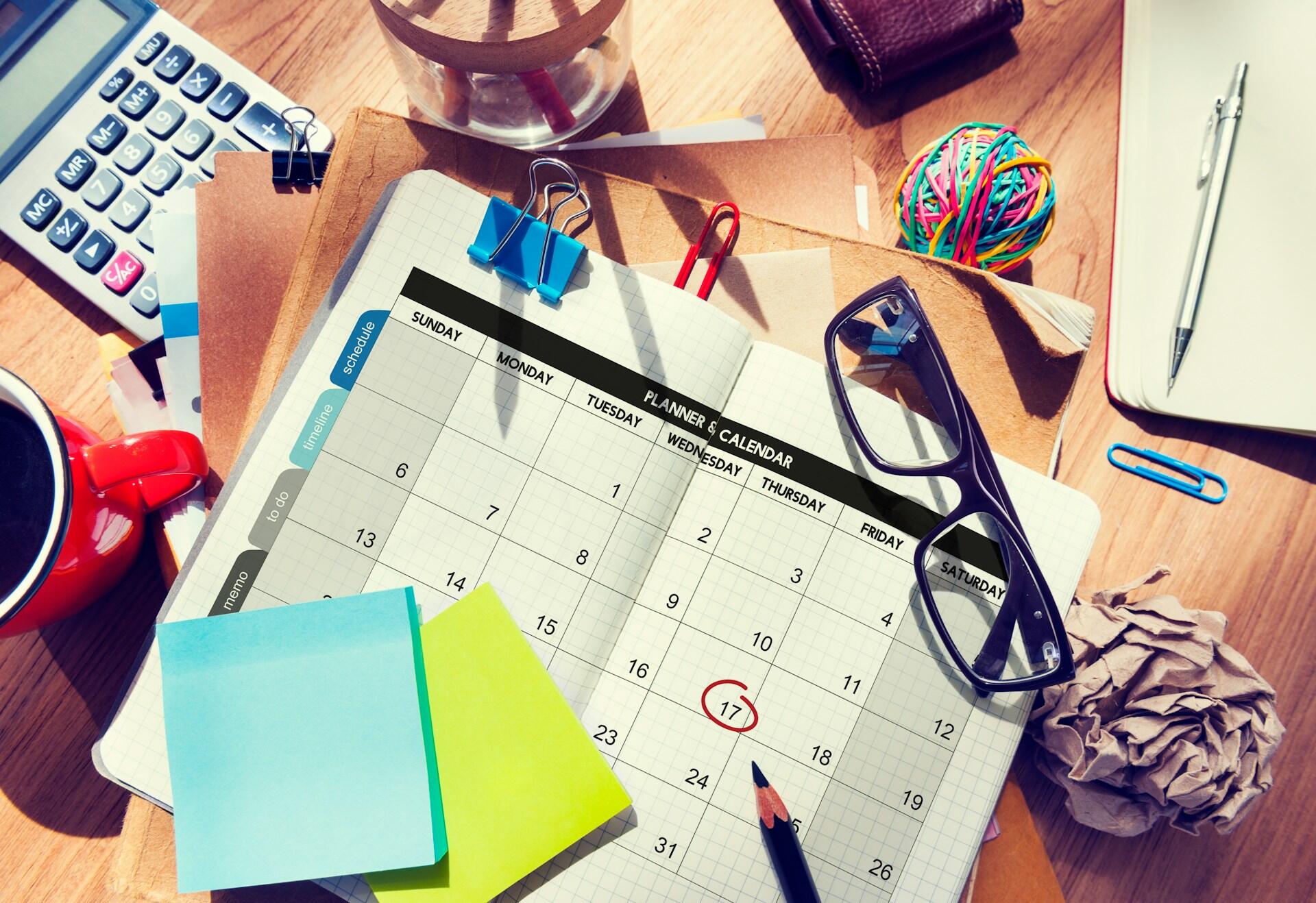 A workstation with a wooden top, featuring an open calendar, black-framed glasses, colourful sticky notes, and a ball of rubber bands.