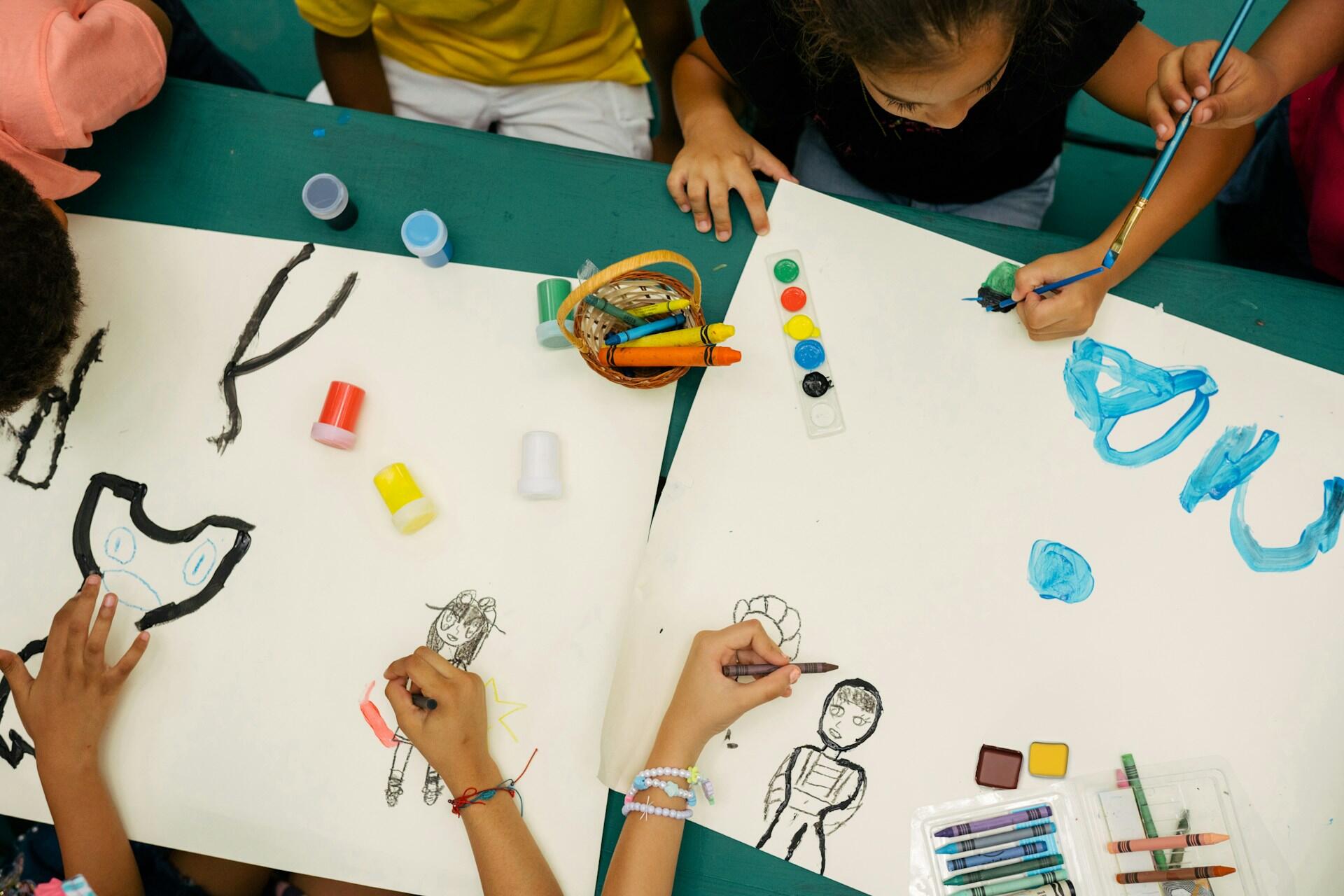 A group of students gathered around a table, creating designs on large white sheets of cardboard.