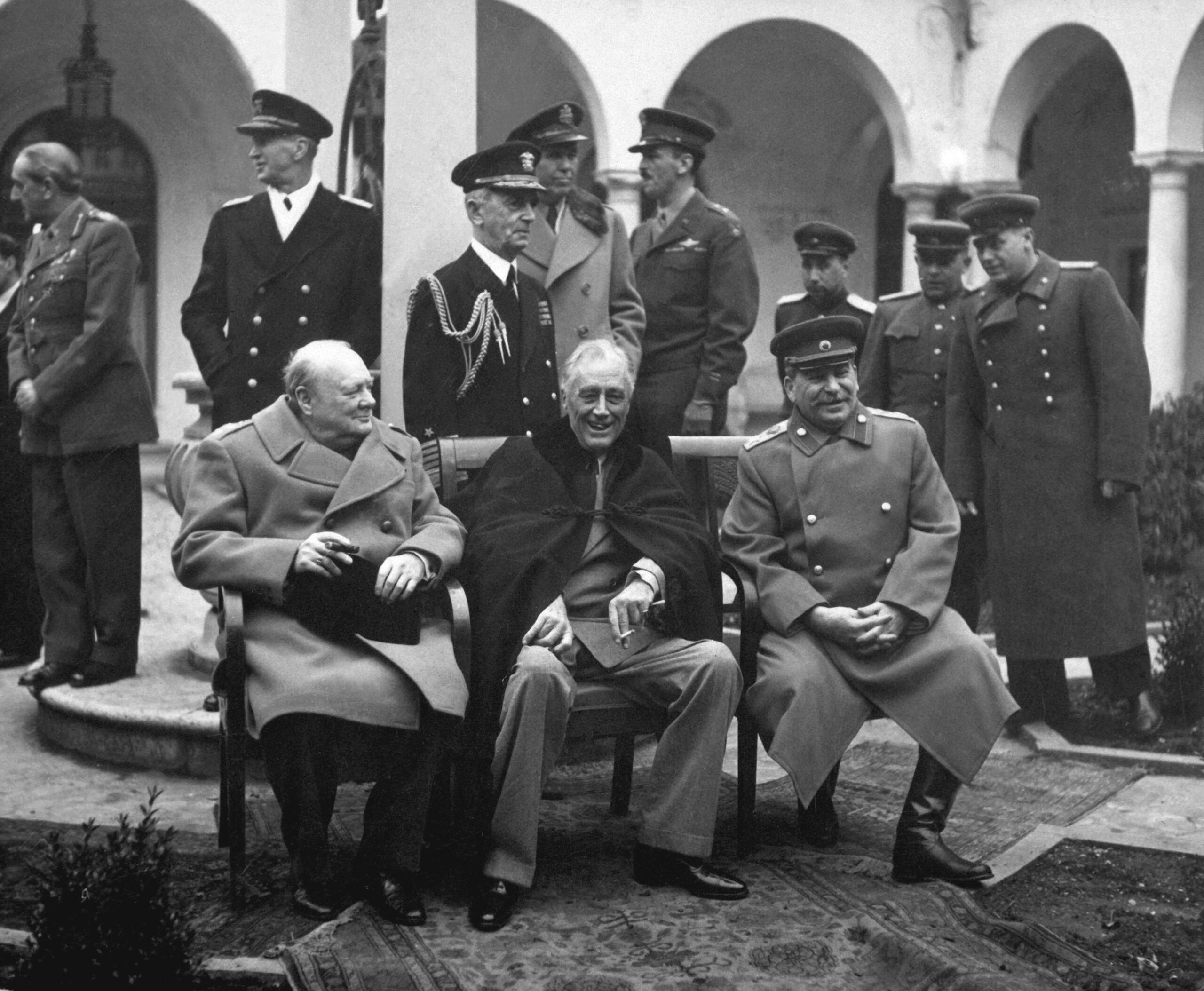 Black-and-white photograph of three Allied leaders seated at the Yalta Conference in February 1945, with military officers standing behind them.