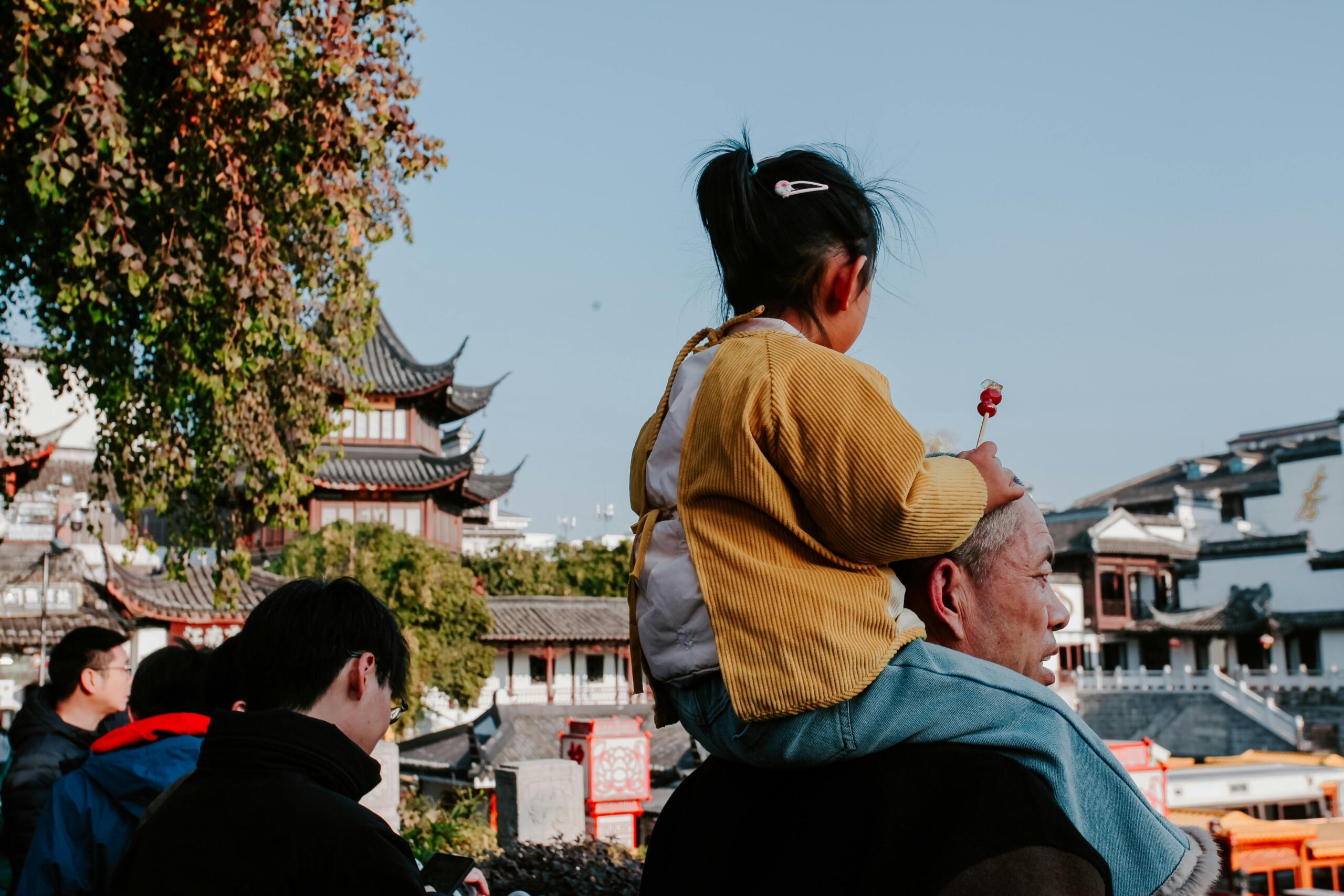 Stariji muškarac nosi dijete na ramenima ispred tradicionalnih kineskih građevina, simbol prenosa znanja o kineskom horoskopu kroz generacije