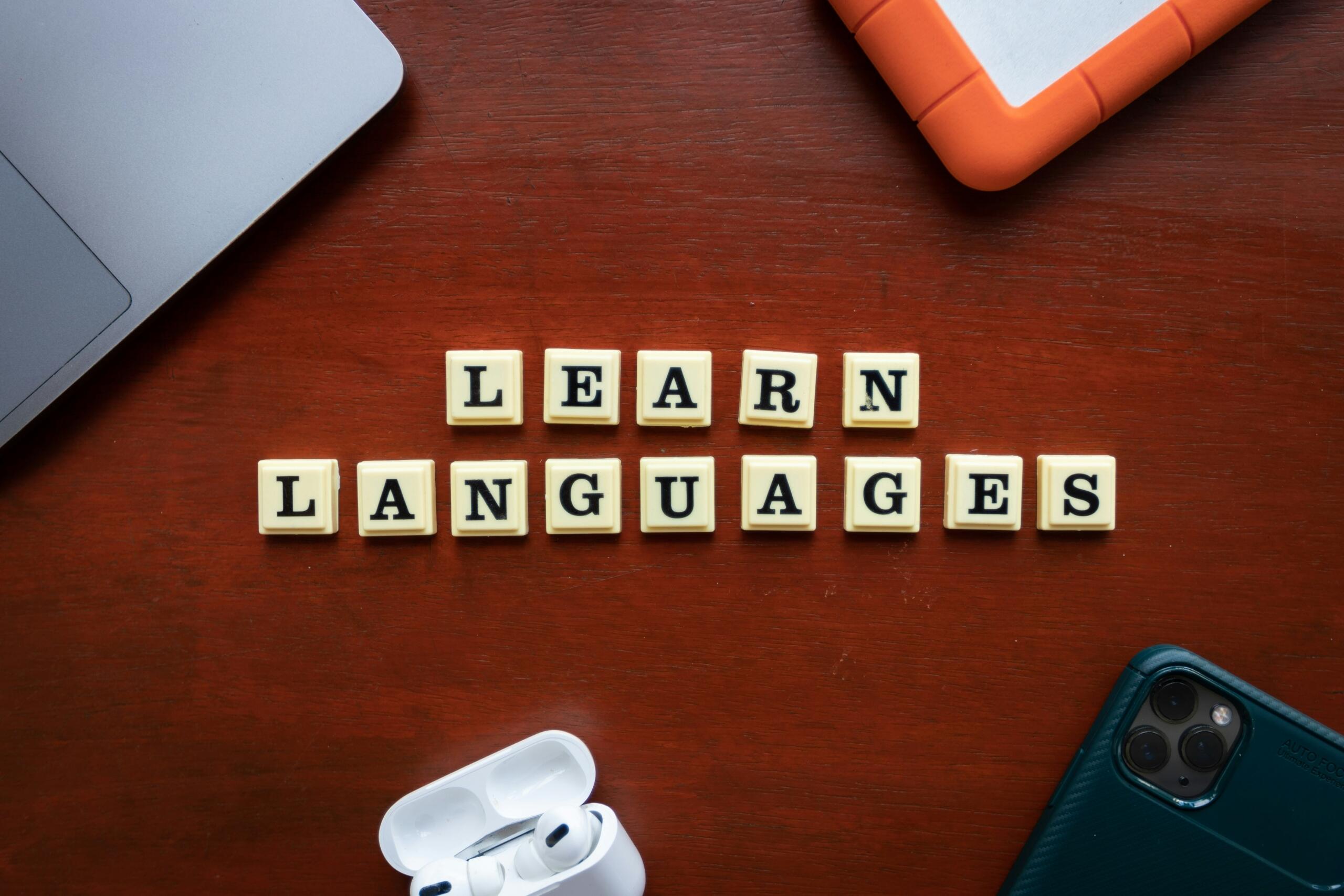 Natpis „Learn Languages” složen od slovnih pločica na drvenom stolu uz laptop, mobitel i slušalice.